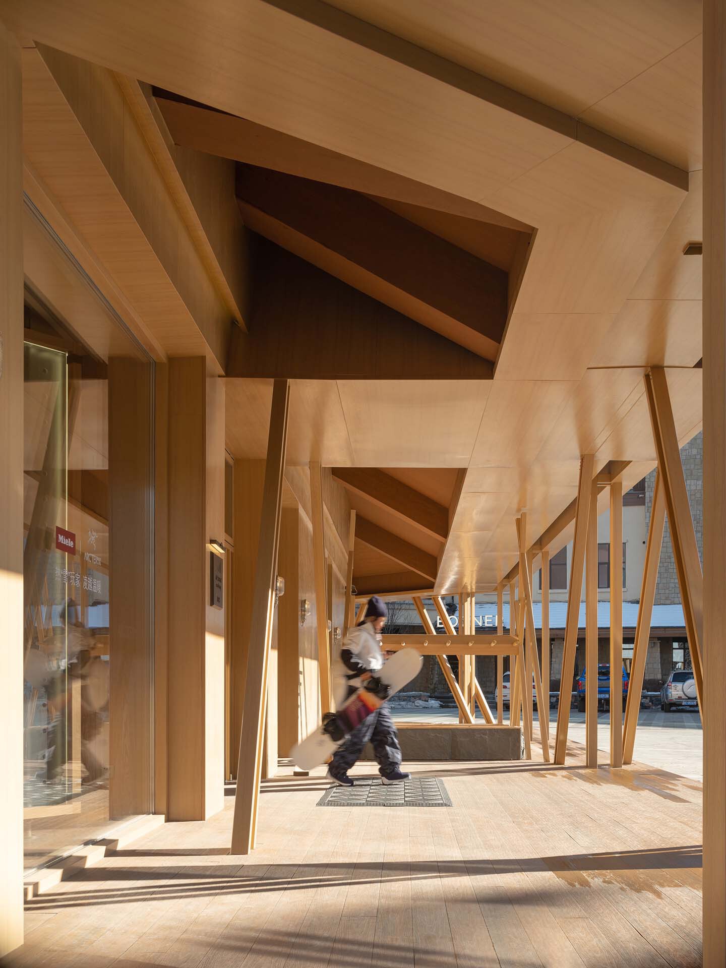 Covered timber colonnade with angled supports and a person walking through dappled sunlight