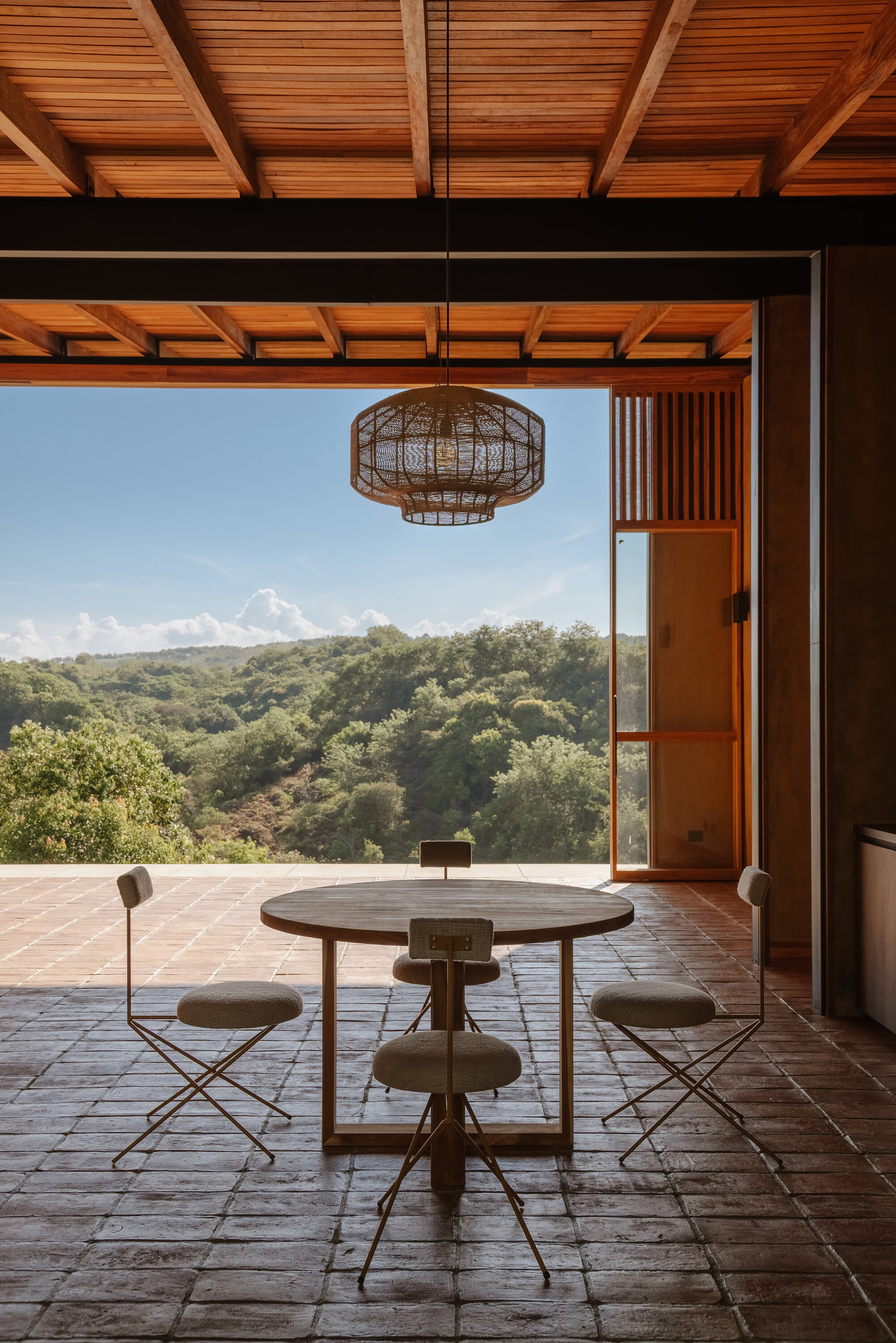 Covered terrace with suspended rattan lantern and round dining table opening to valley view