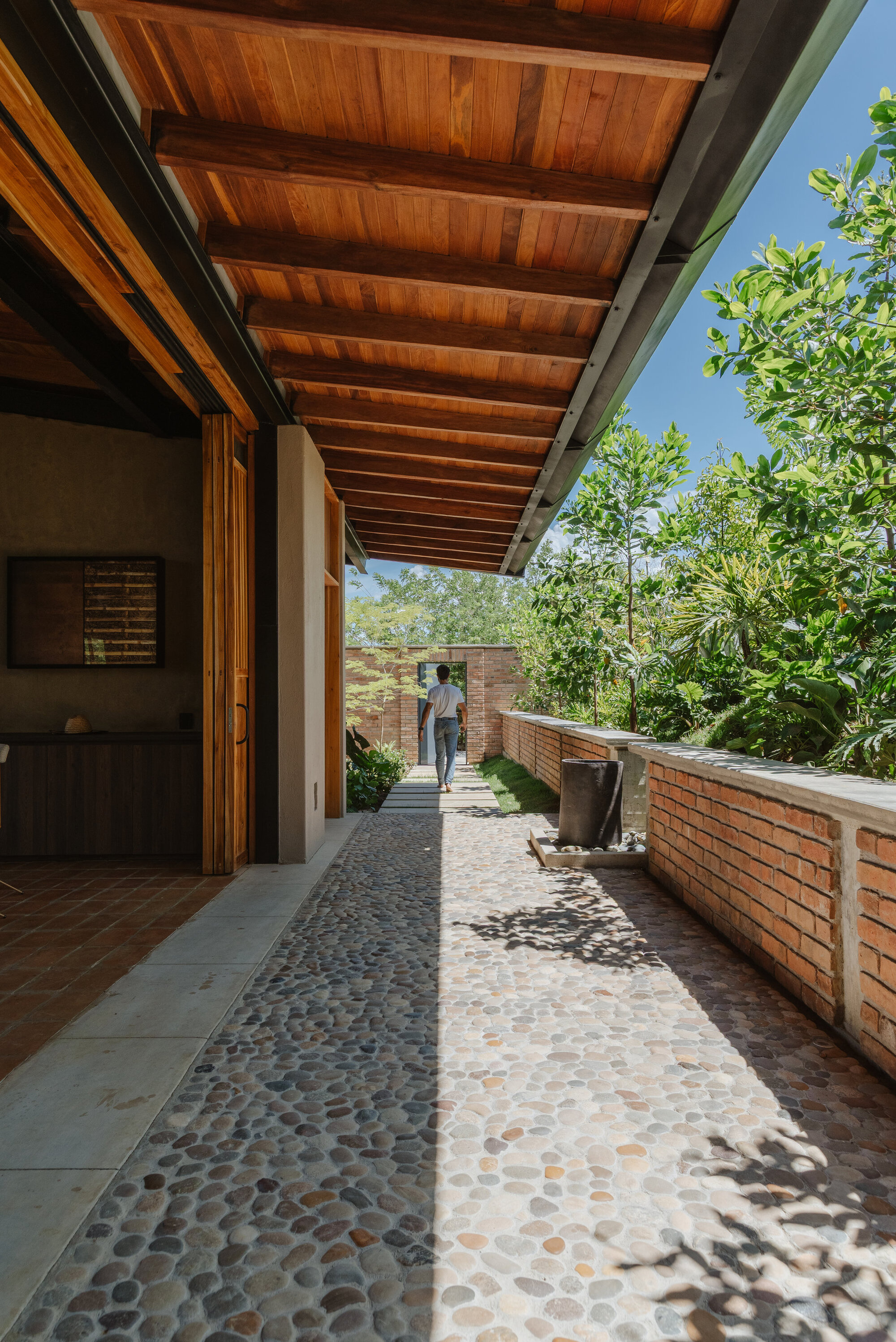 Covered walkway with river stone pathway and timber overhang alongside planted bed with figure passing through