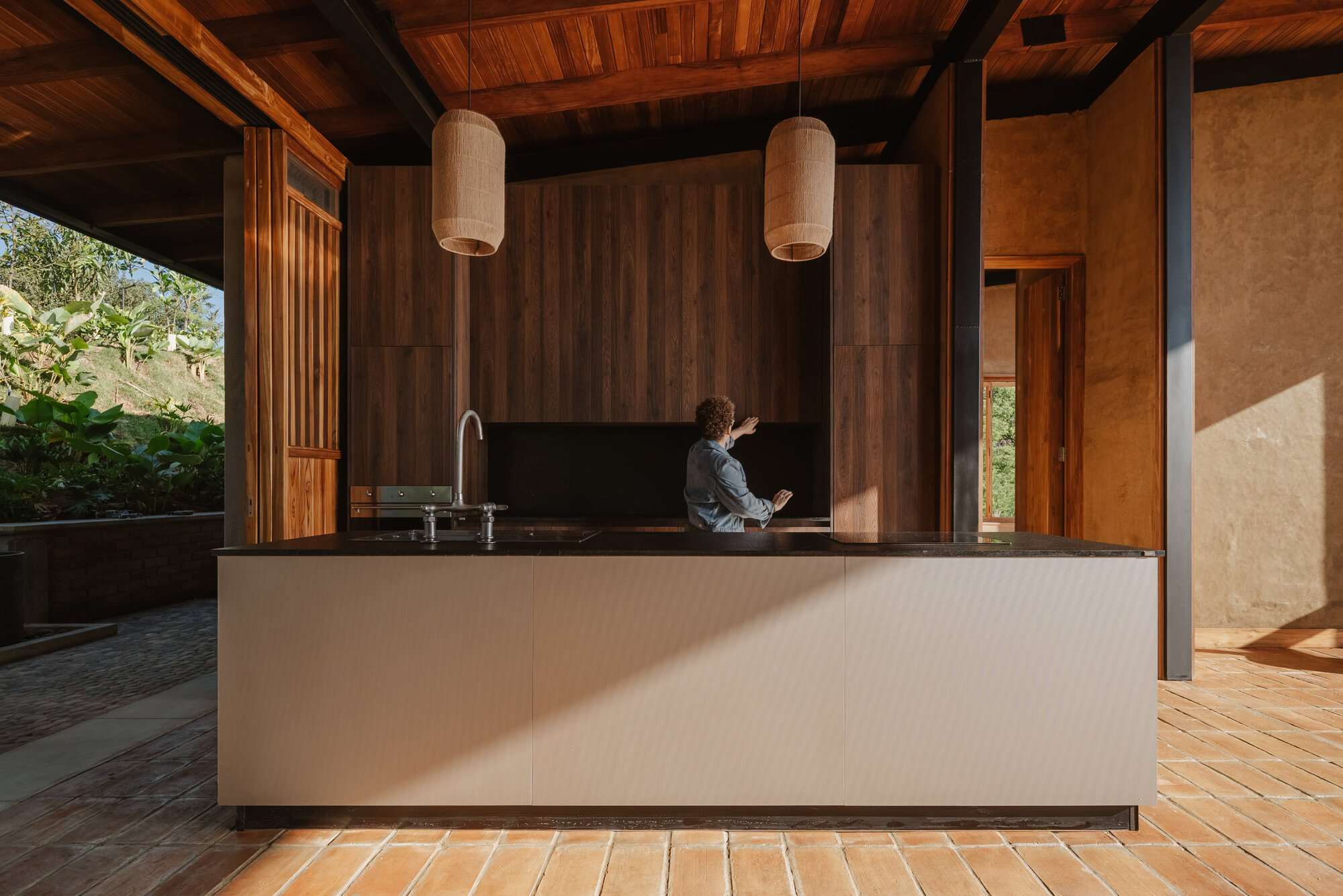 Kitchen island with black countertop under timber ceiling and woven pendant lights with terracotta floor tiles