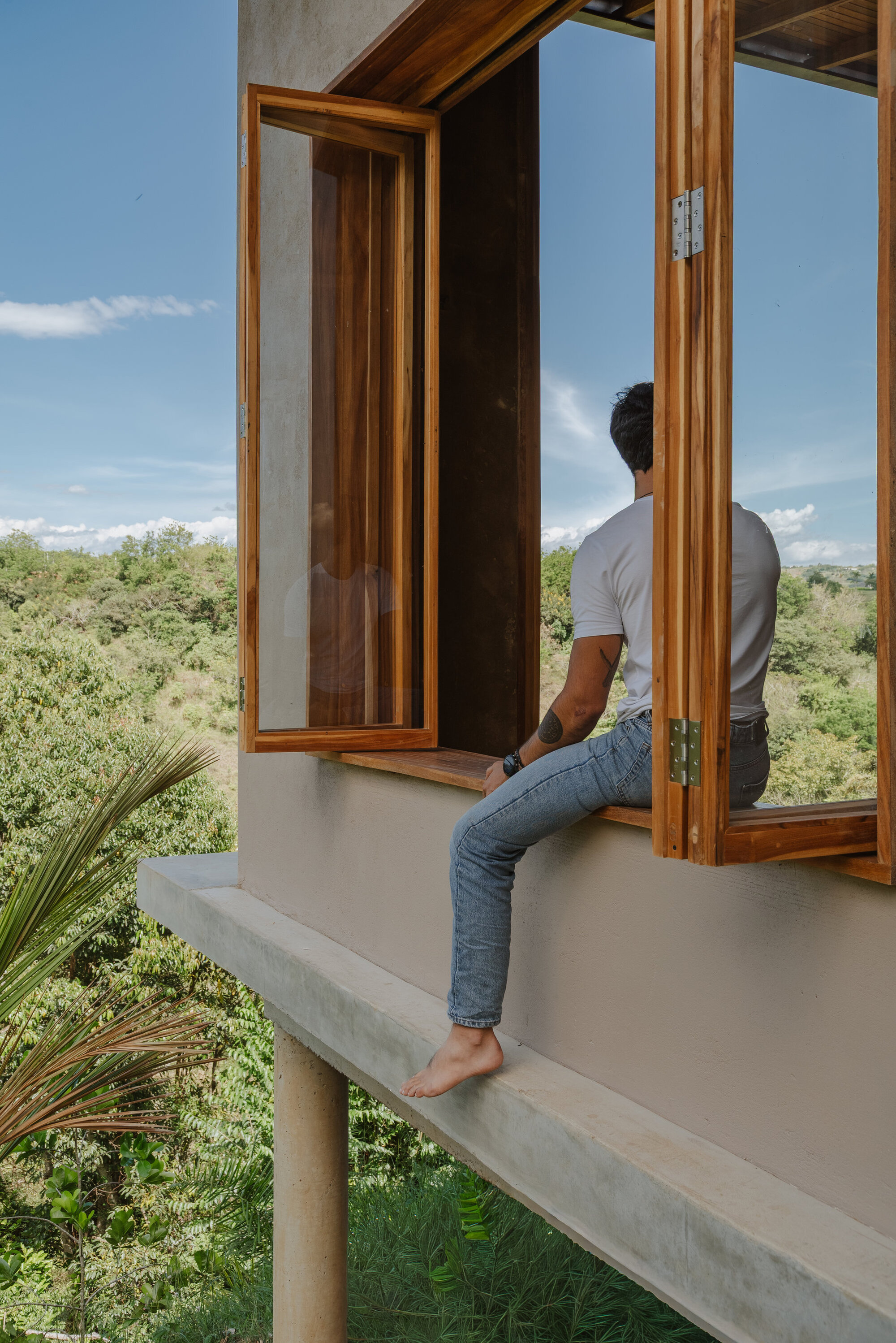 Person seated on cantilevered concrete sill at timber casement window overlooking valley landscape