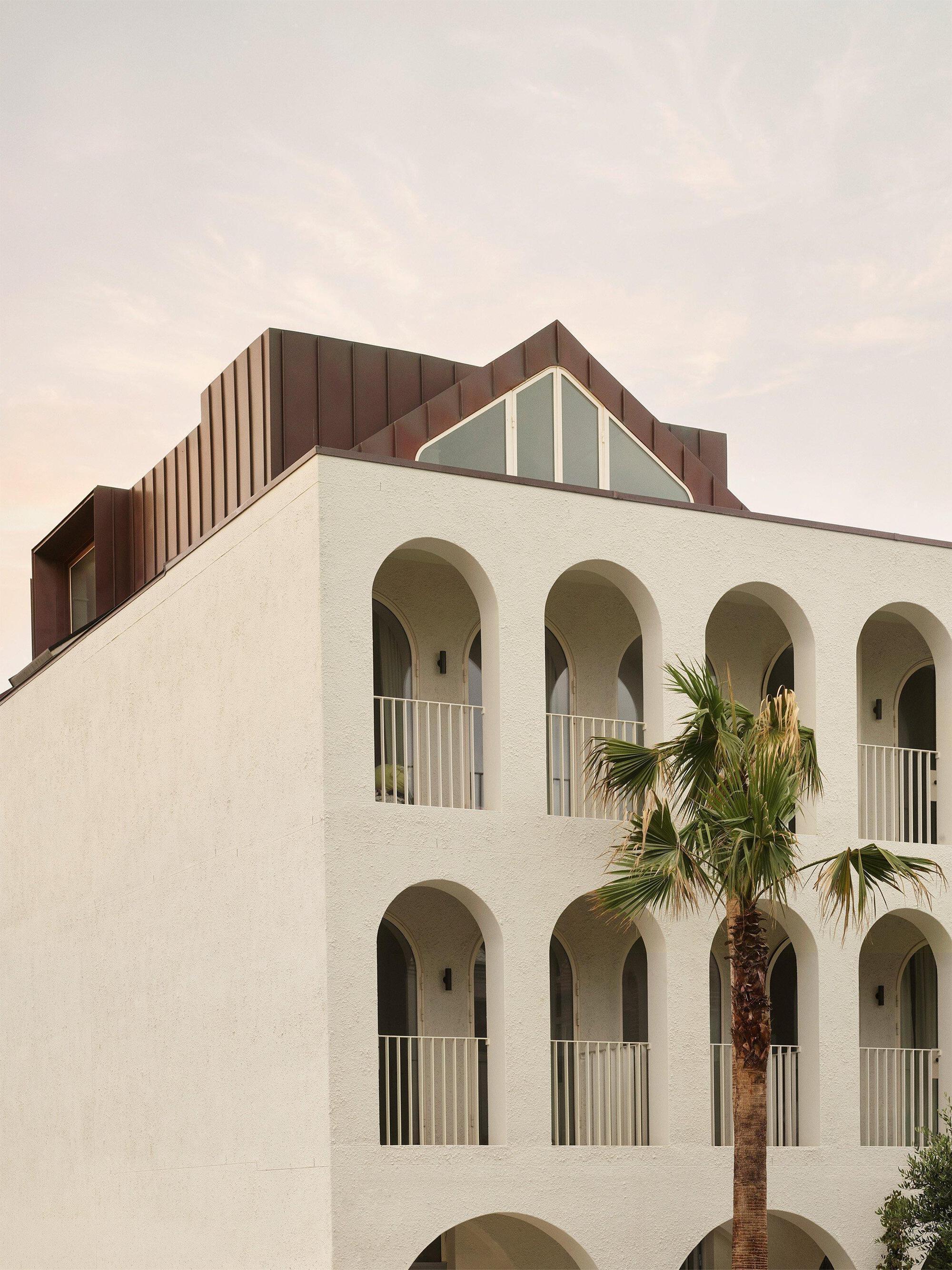 White painted facade with arched balconies and a brown metal-clad upper volume against a hazy sky