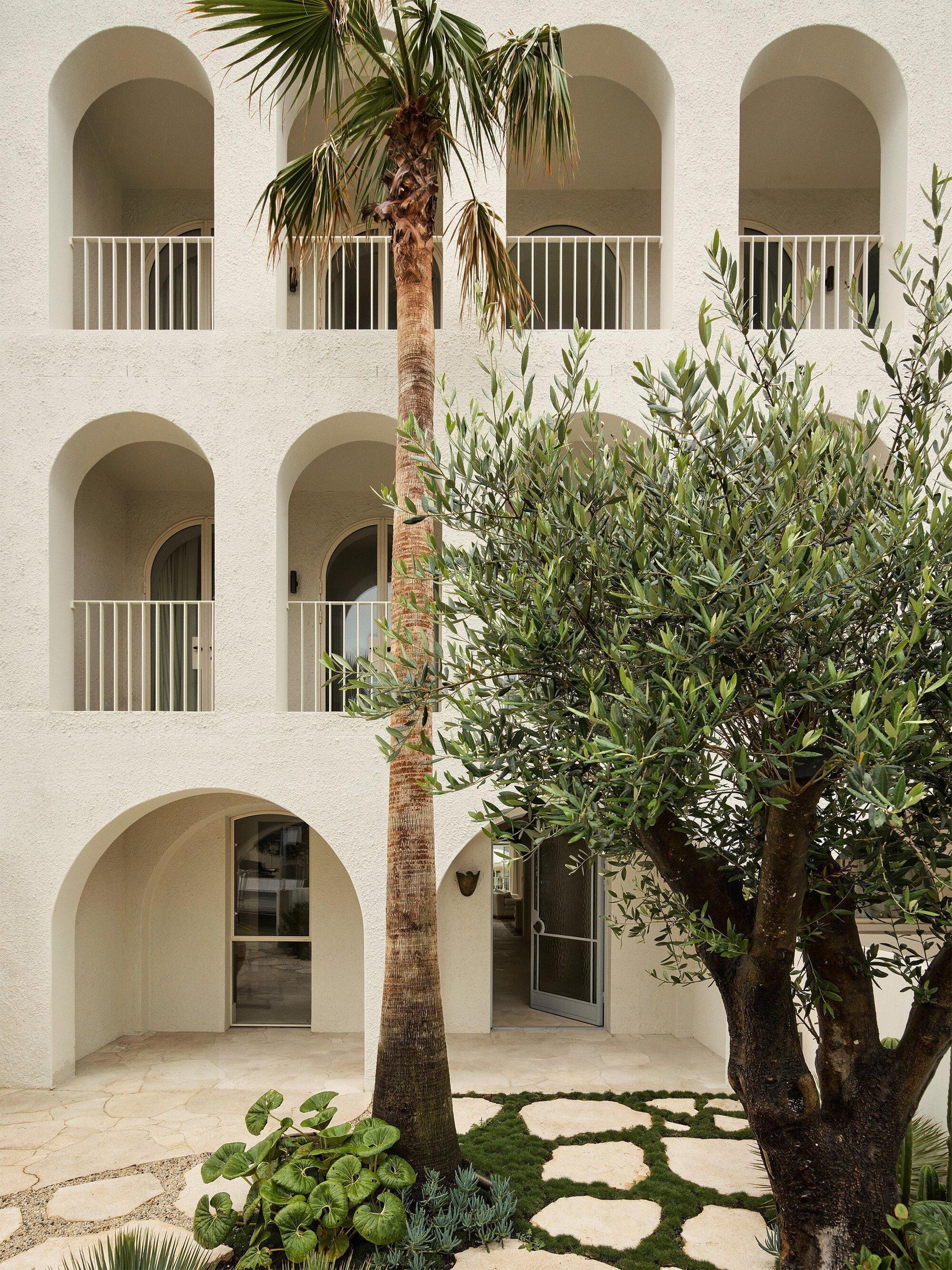 Courtyard view of arched loggias with white railings and planted bed with palm and olive trees