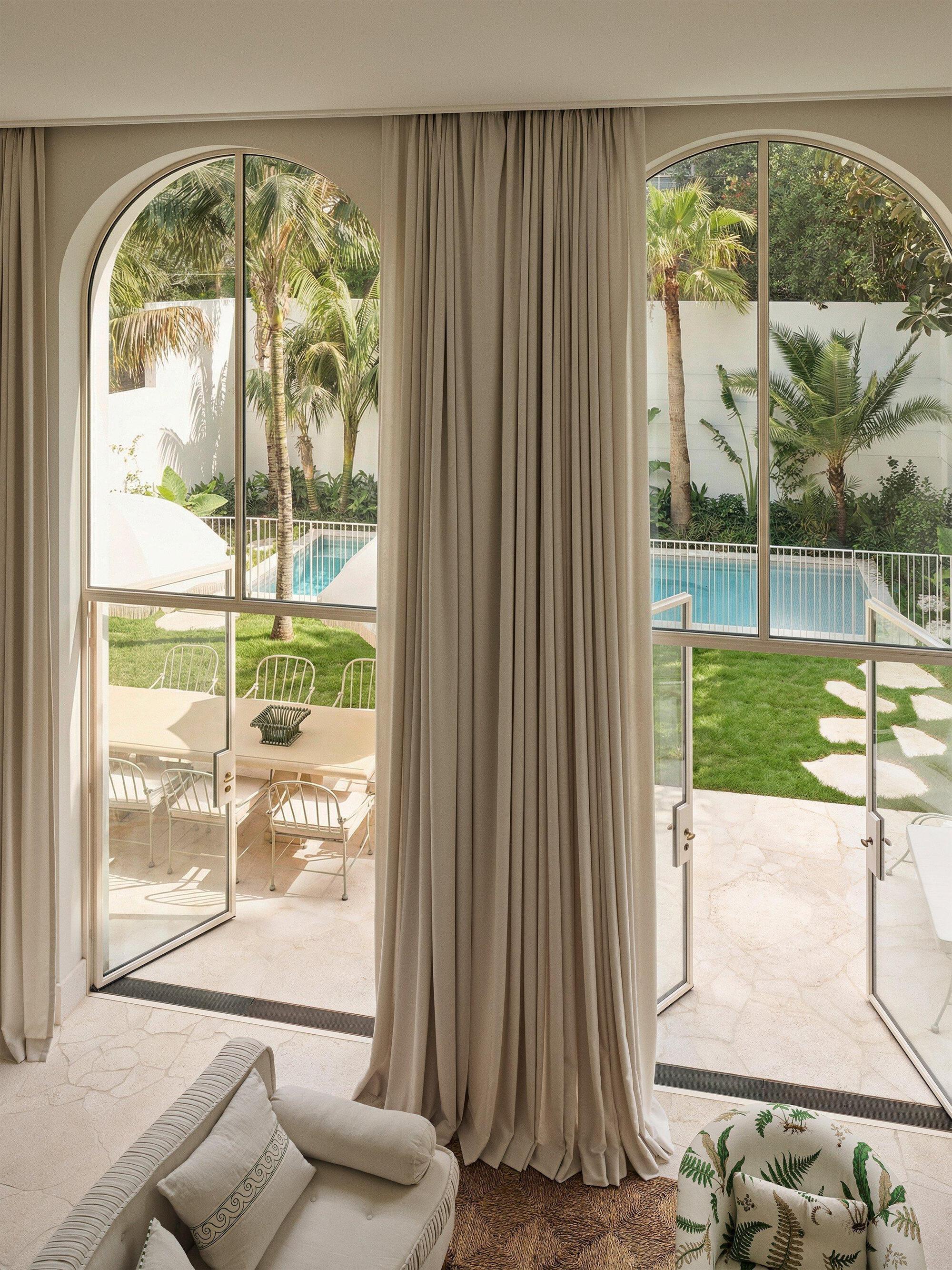 Interior view through arched glass doors with linen curtains overlooking a pool and lawn with palms