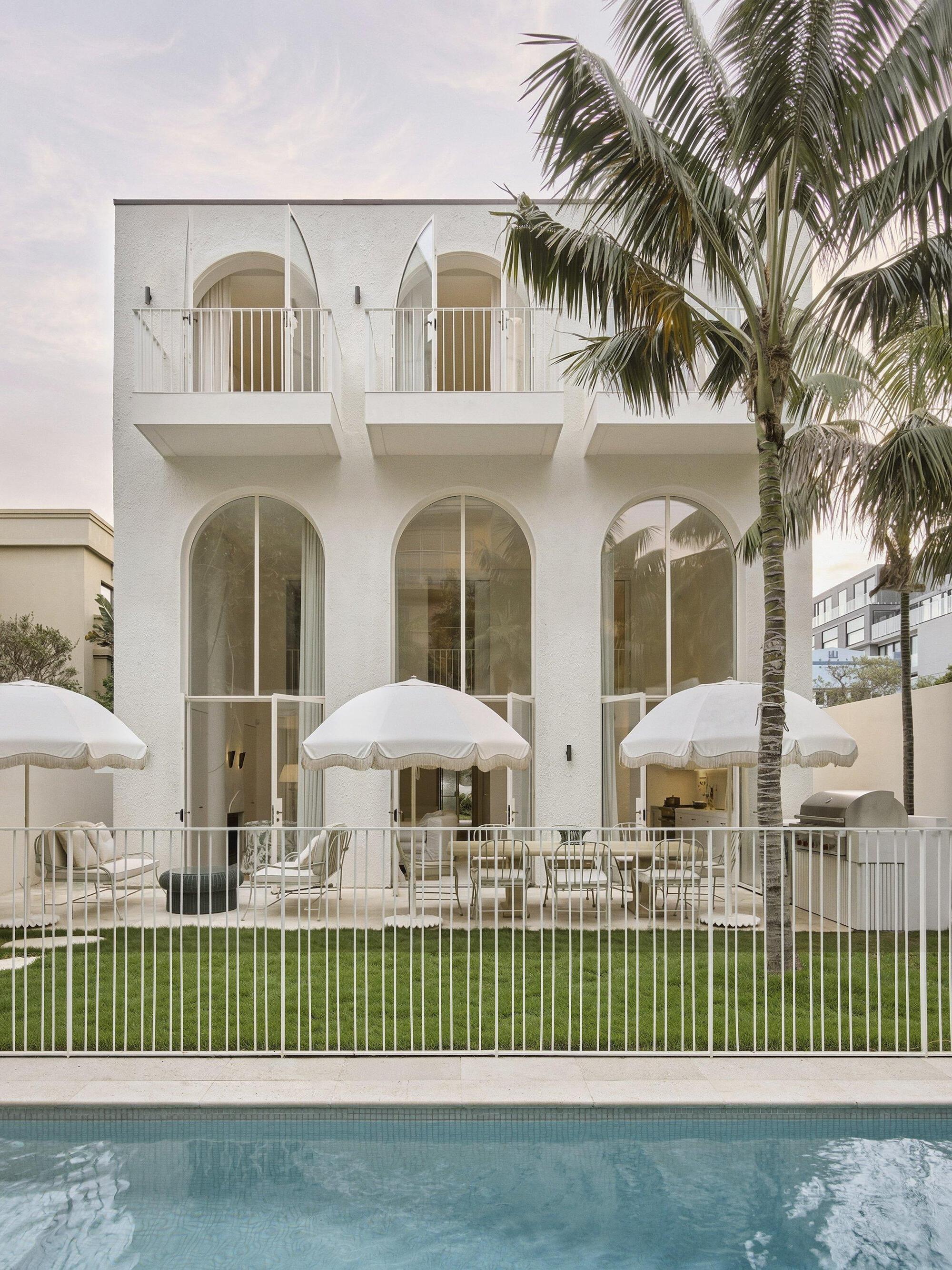 White stucco facade with arched openings and upper balconies beside a pool and palm trees