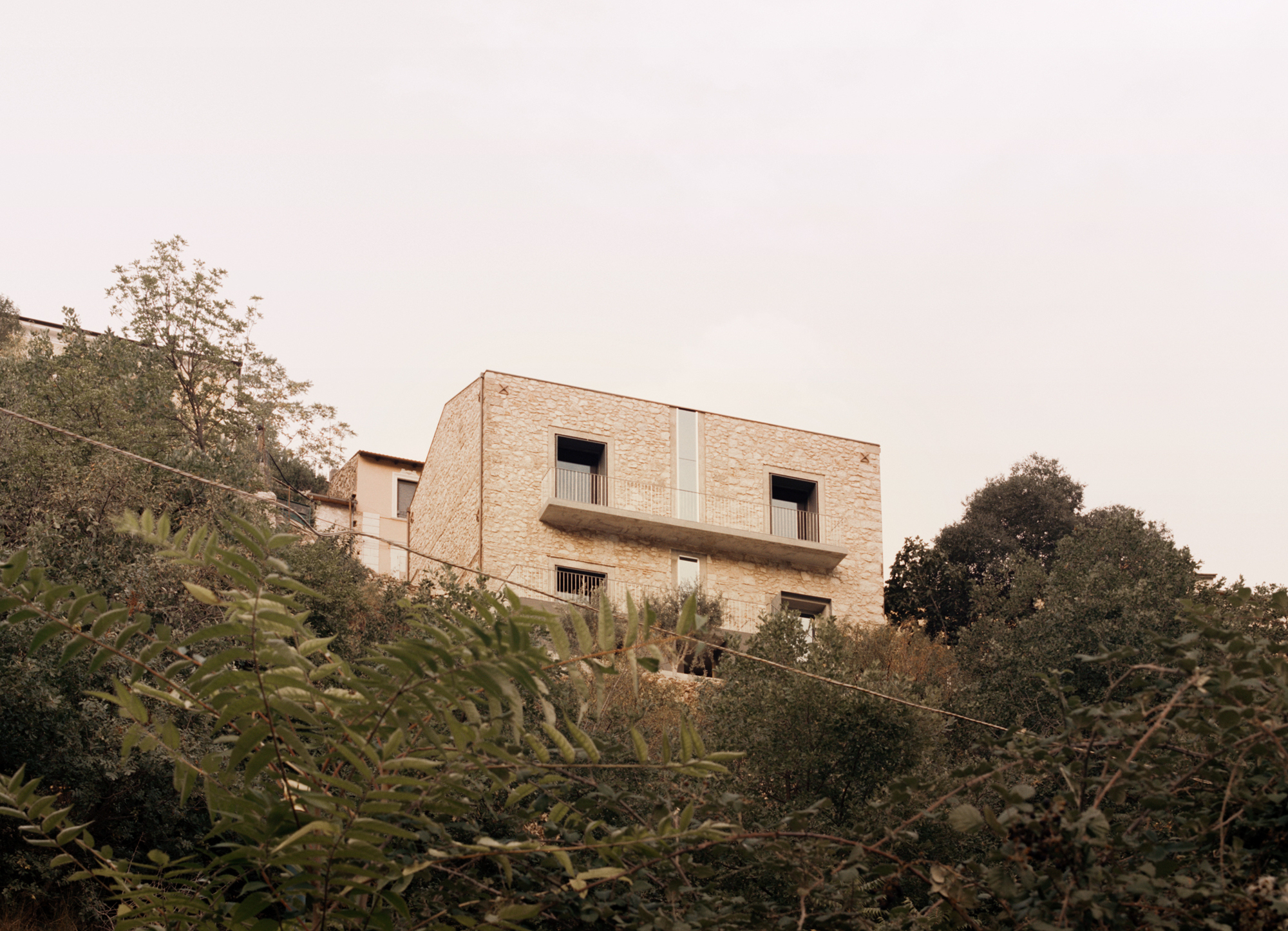 Stacked brick volume rising from a hillside with dense vegetation and overcast sky