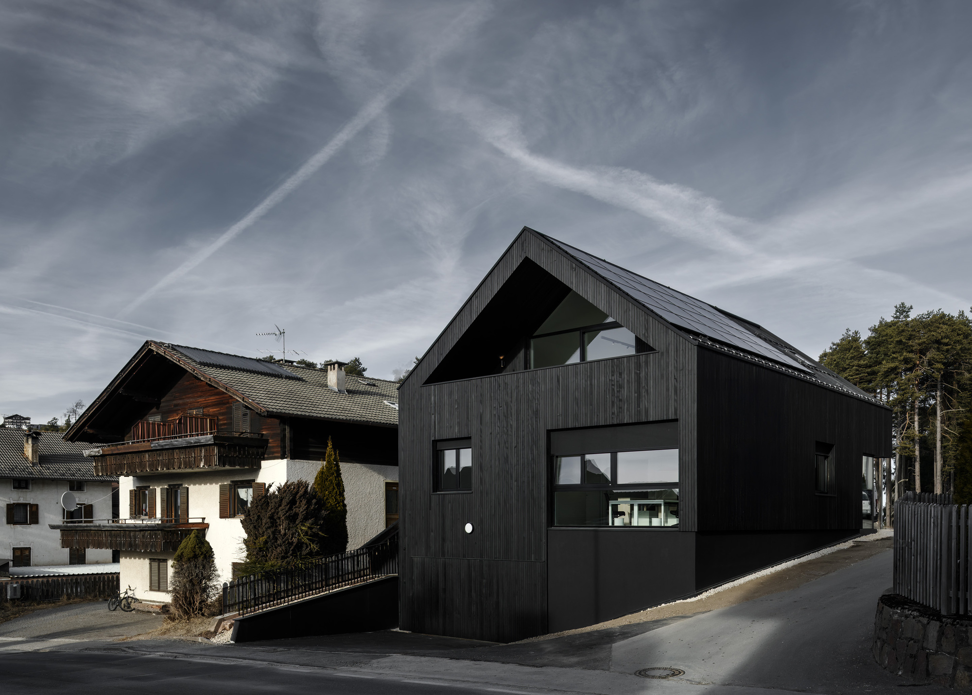 Black vertical timber cladding on a gabled volume adjacent to a traditional alpine house under overcast skies