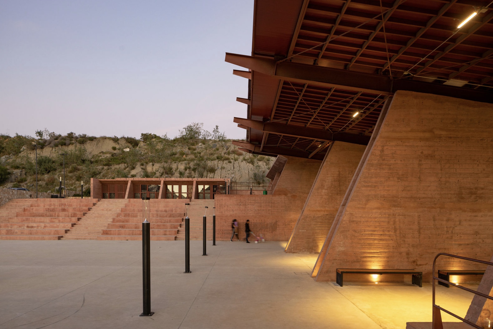 Cantilevered timber-framed canopy over a concrete plaza with stepped seating at dusk