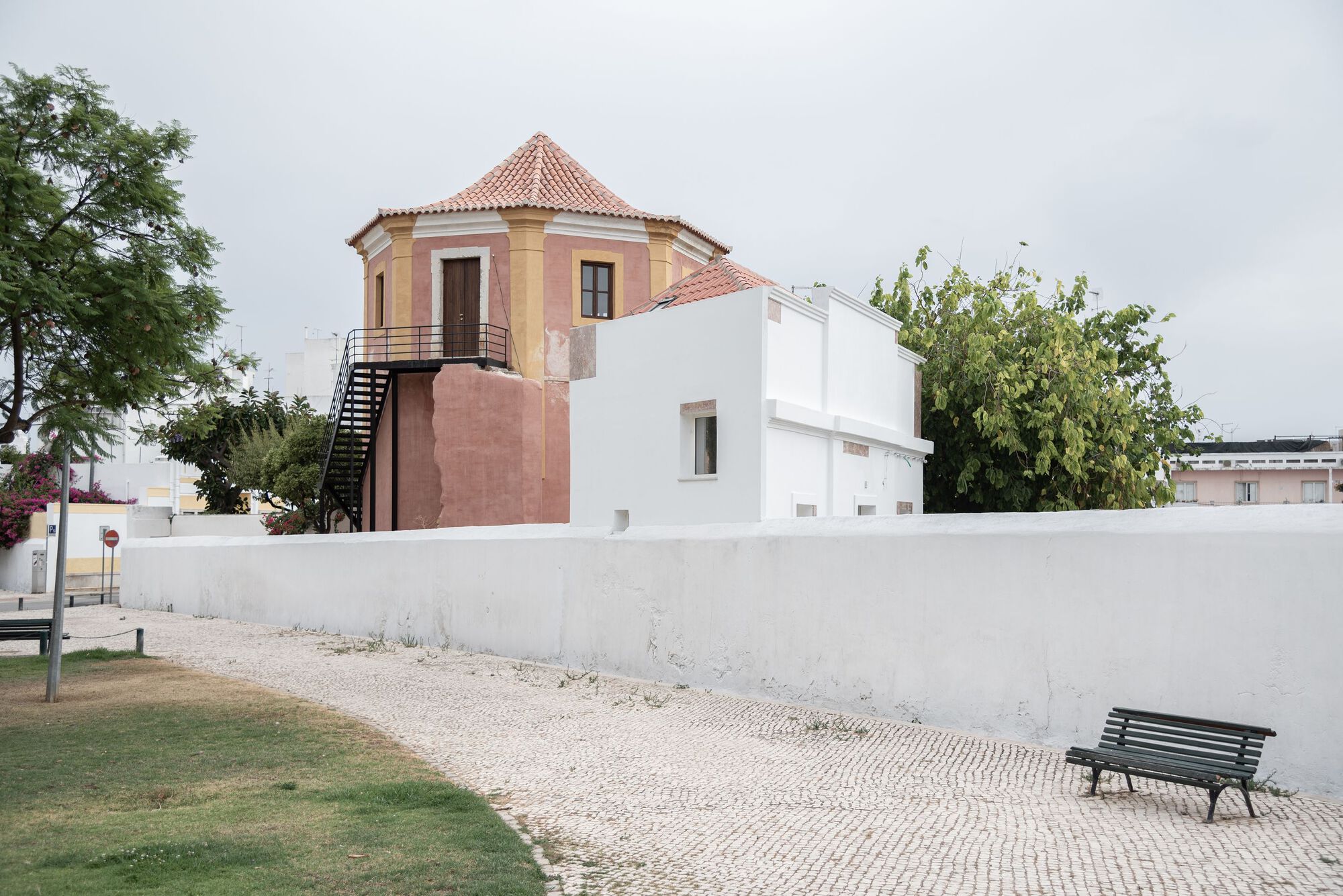 Octagonal tower with terracotta roof tiles adjacent to renovated white volumes under overcast skies