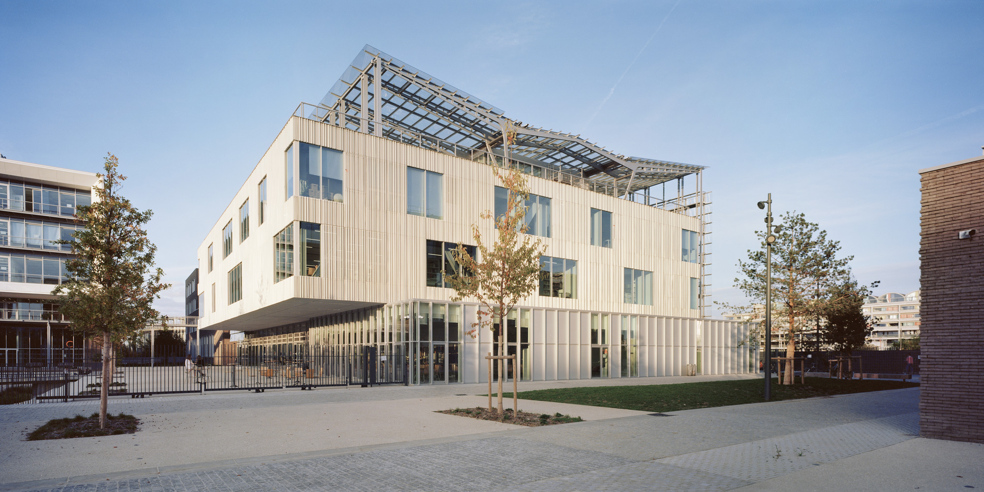 Street facade with vertical metal cladding and a translucent glazed roof structure under afternoon sun