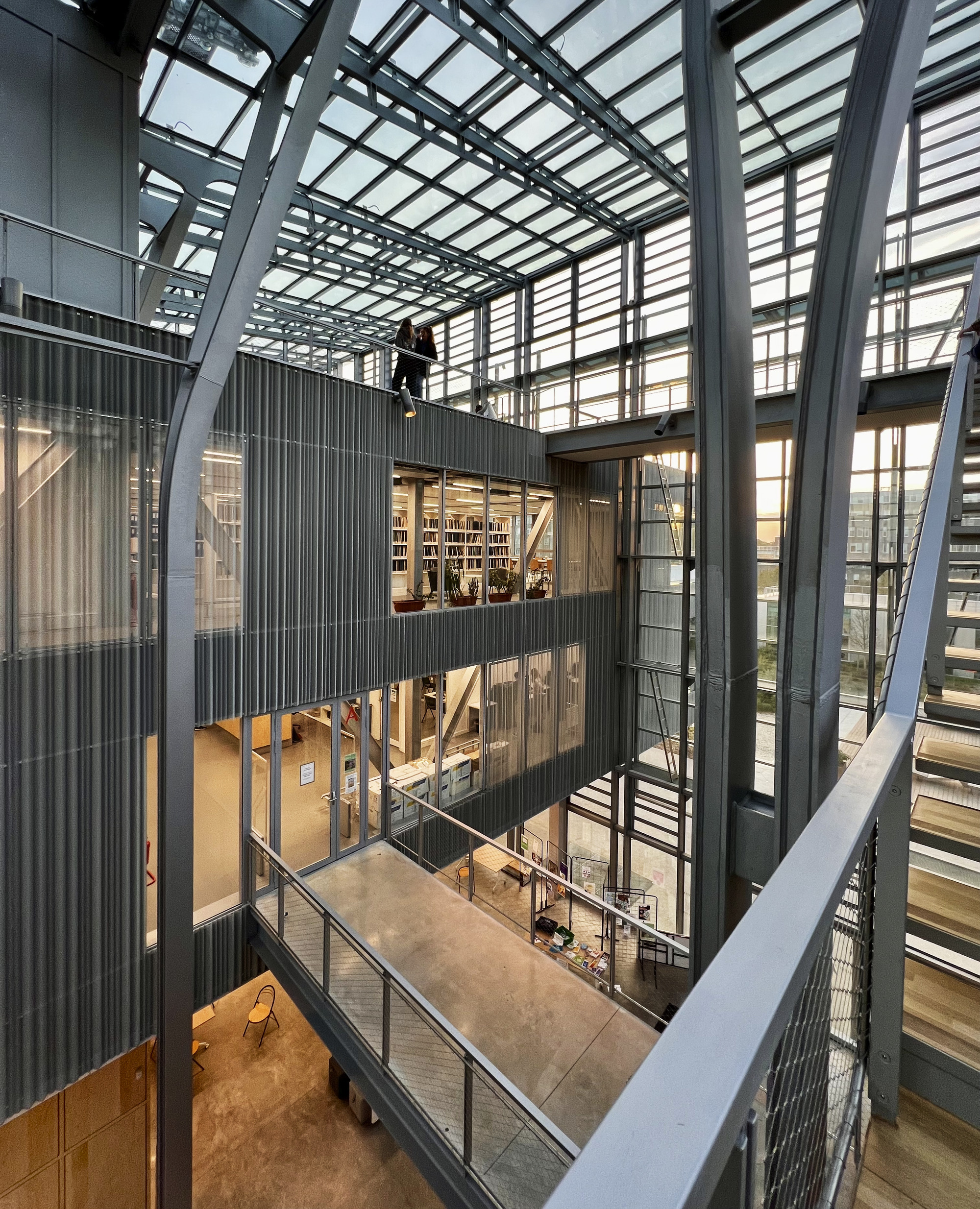 Multi-story atrium with corrugated metal volumes suspended between exposed steel structural framework and glass ceiling