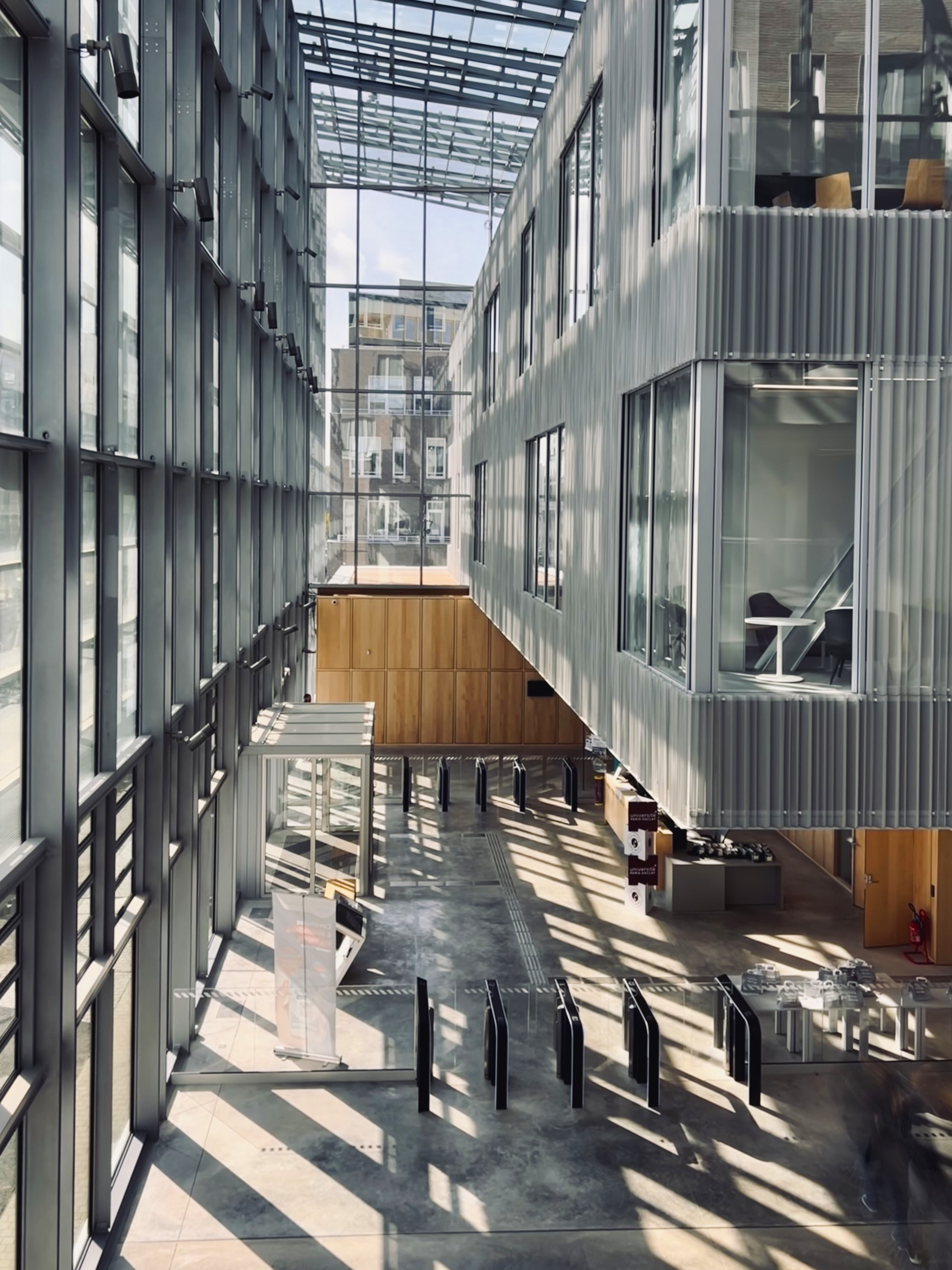 Glazed atrium corridor with steel and glass roof casting shadows across the concrete floor at midday
