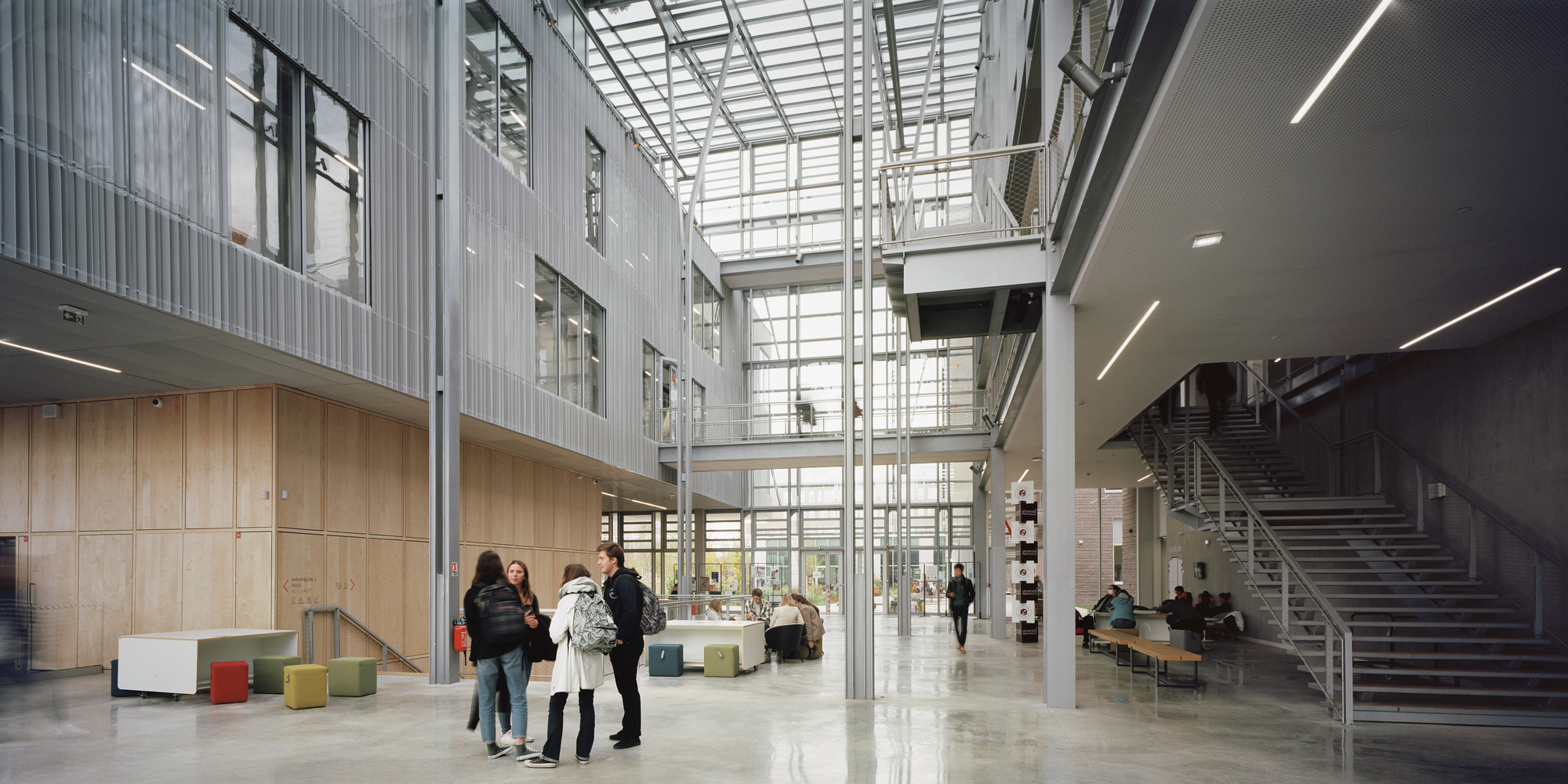 Multi-level lobby with glass roof, metal staircase, and groups of visitors gathered near wood paneled walls
