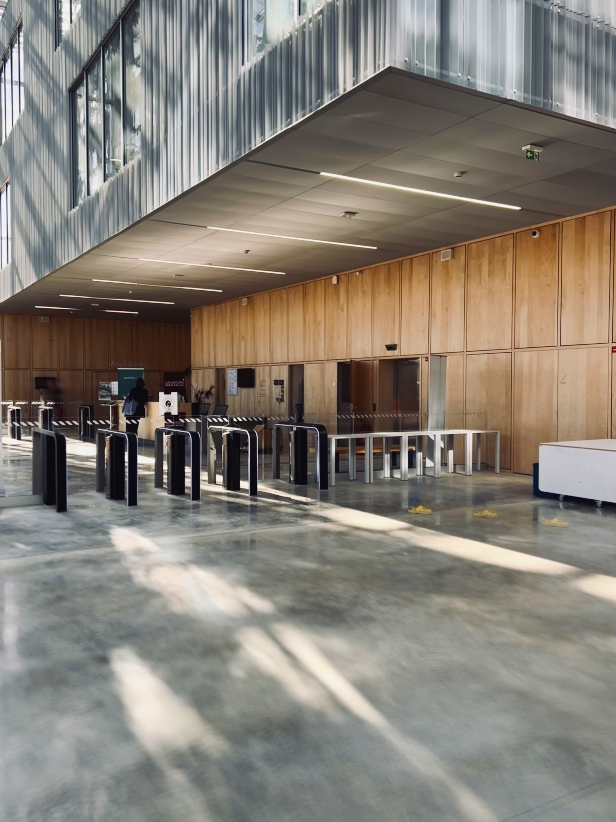 Entry lobby showing timber wall panels and turnstile gates beneath a concrete soffit with linear light fixtures