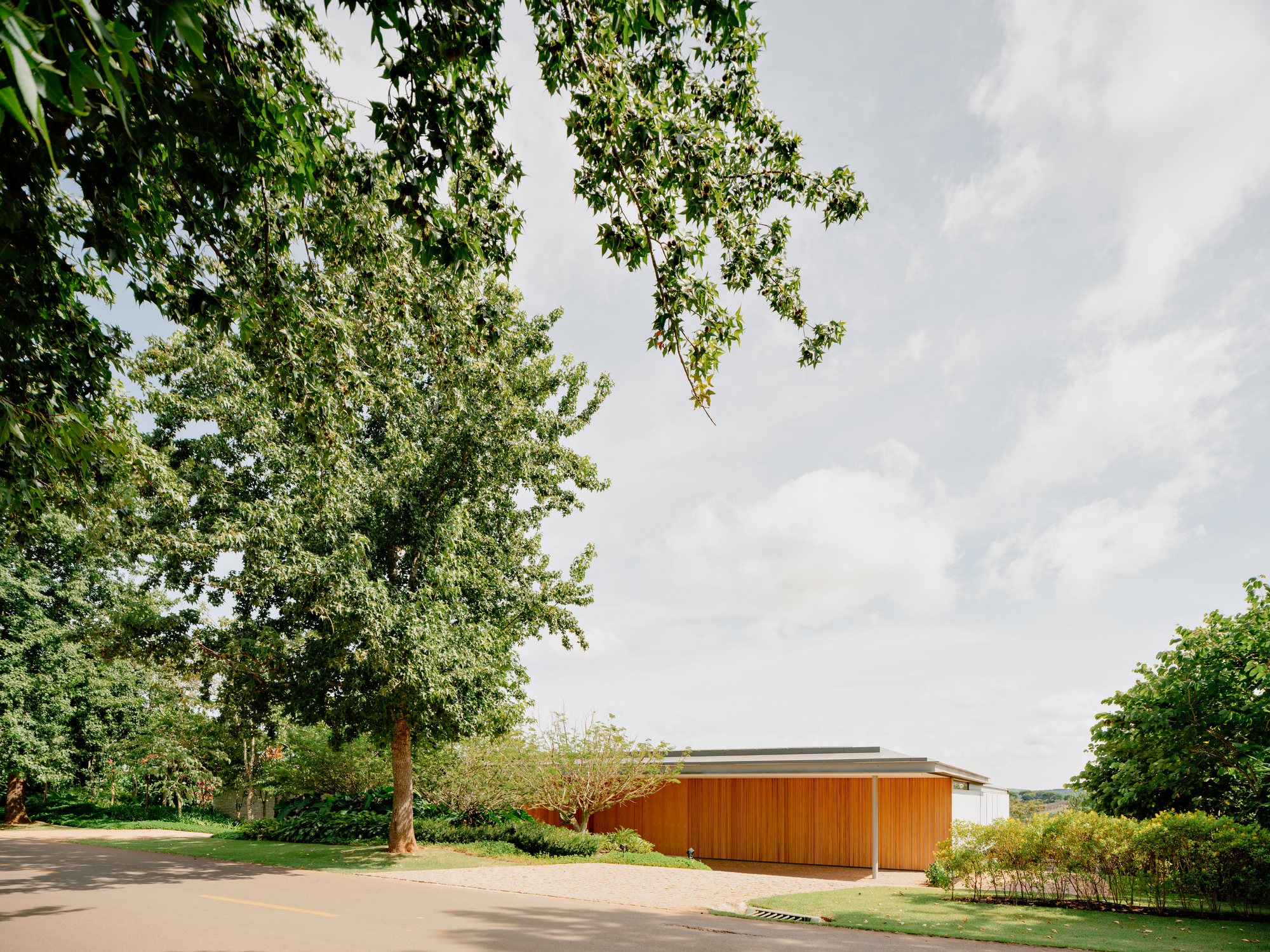 Horizontal timber facade and flat roof cantilevering over a gravel drive edged by lawn and trees