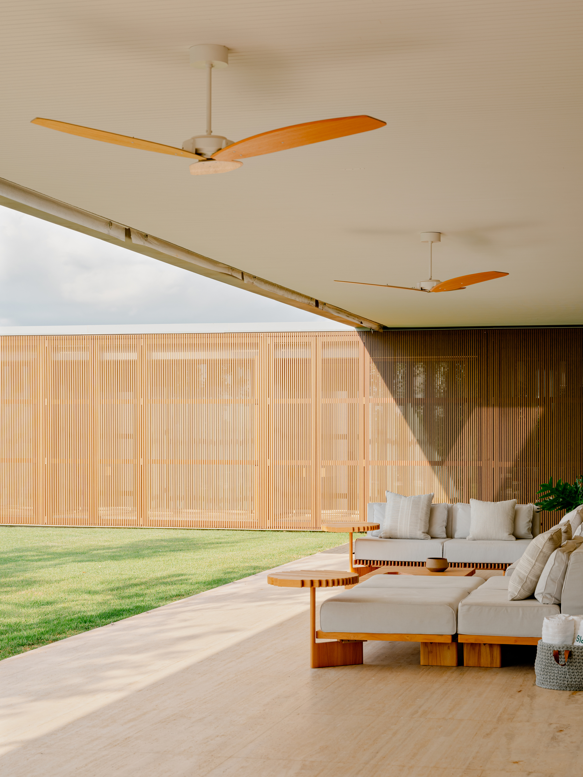 Timber slatted screen wall beneath a ceiling with two three-bladed fans and afternoon sunlight on the floor
