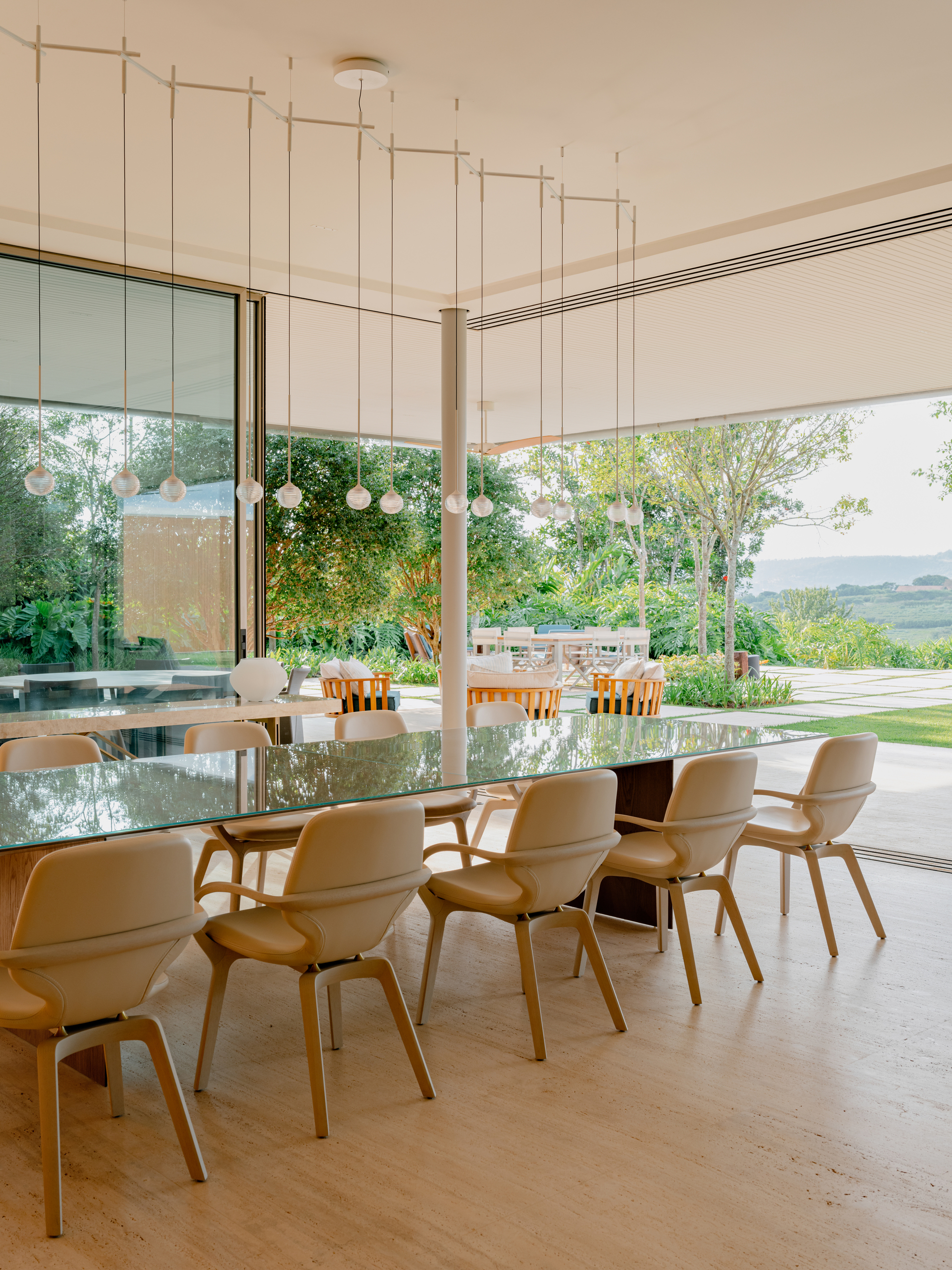 Dining table with curved-back chairs under a suspended ceiling grid facing floor-to-ceiling glazing and garden beyond