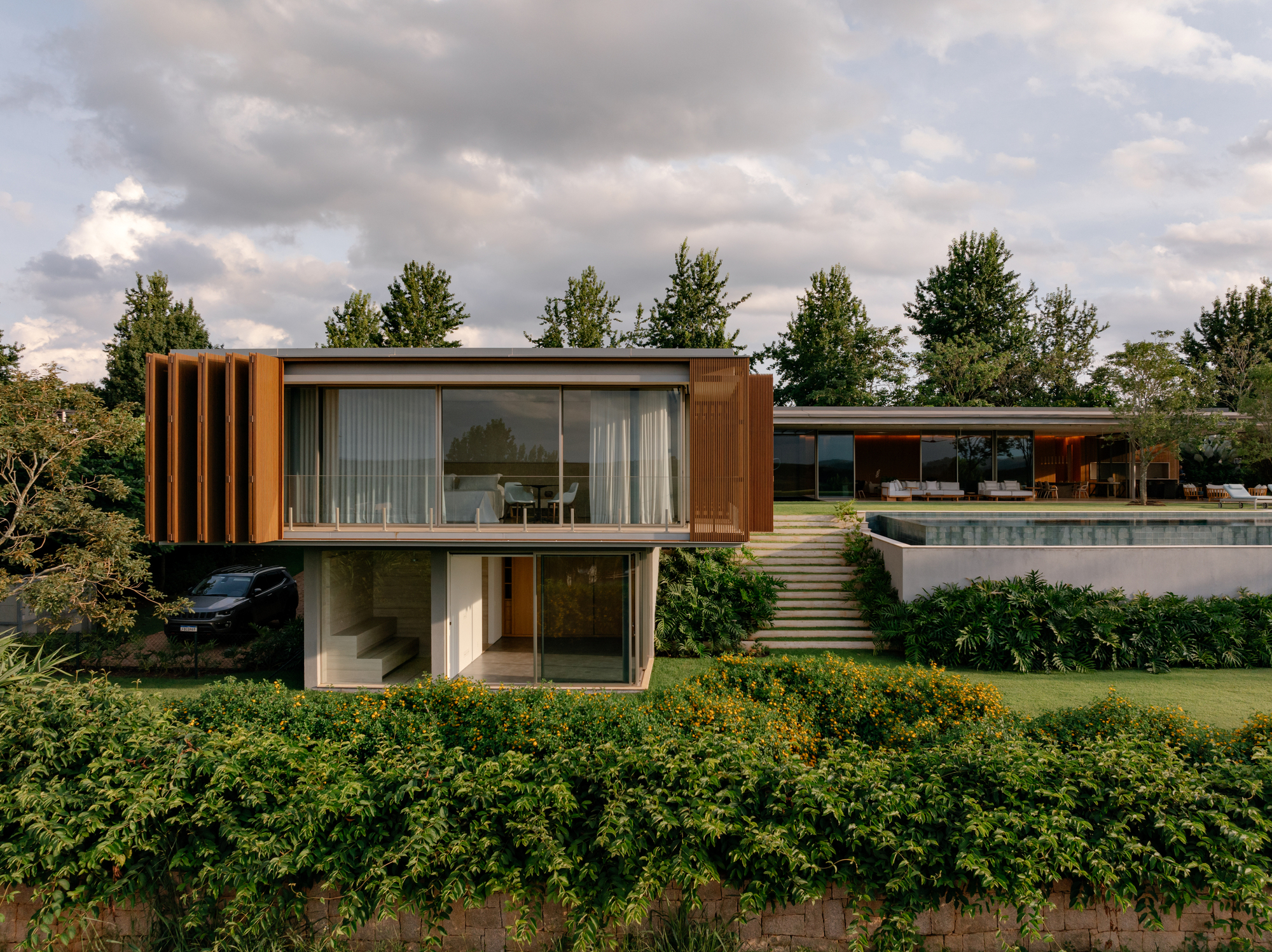 Two-story volume with vertical timber slats and full-height glazing overlooking a garden and pool under cloudy skies