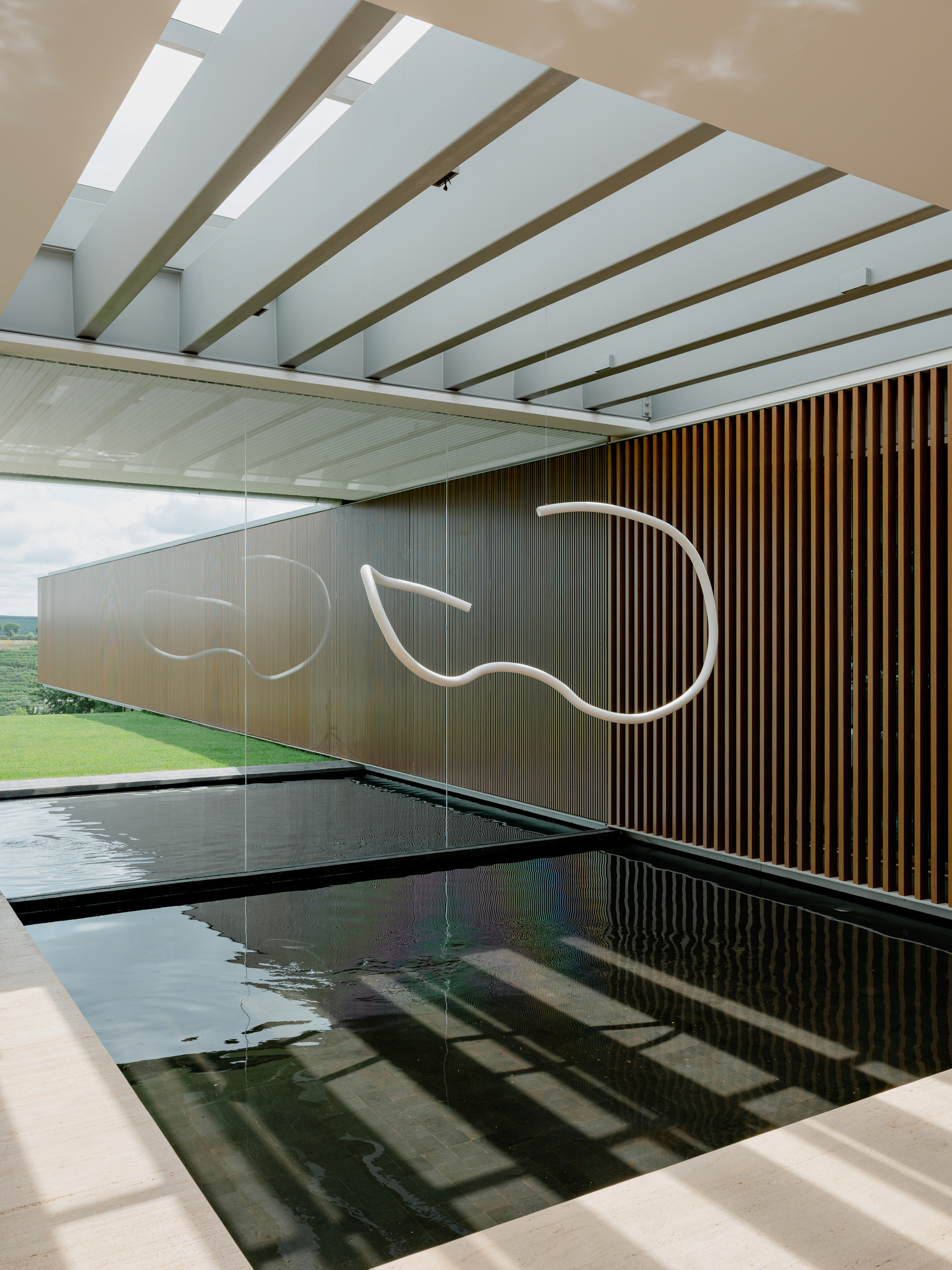 Indoor lap pool with dark tile lining beneath exposed timber beams and skylights running the length