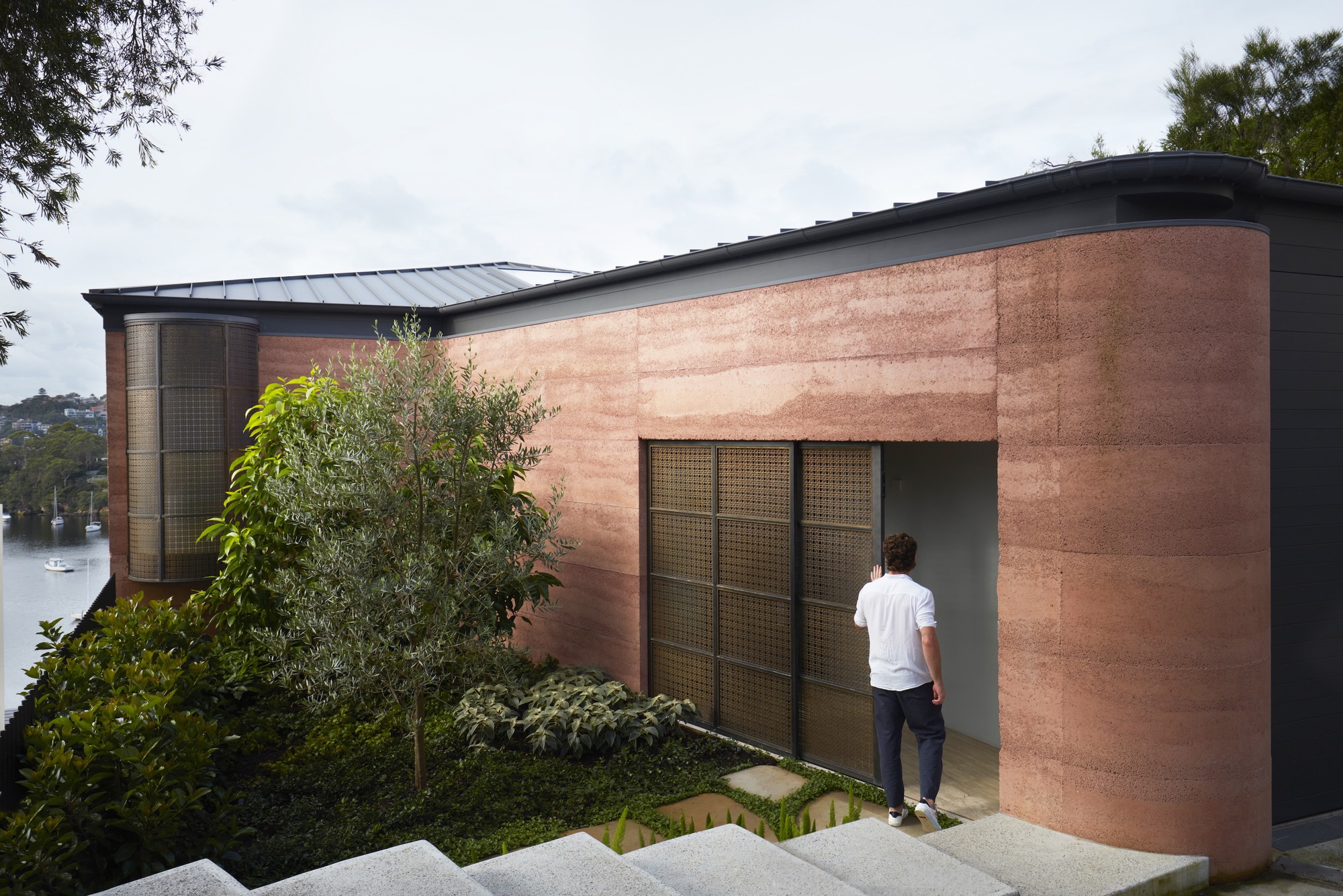 Curved rammed earth wall with recessed glass door and person entering beneath overcast sky