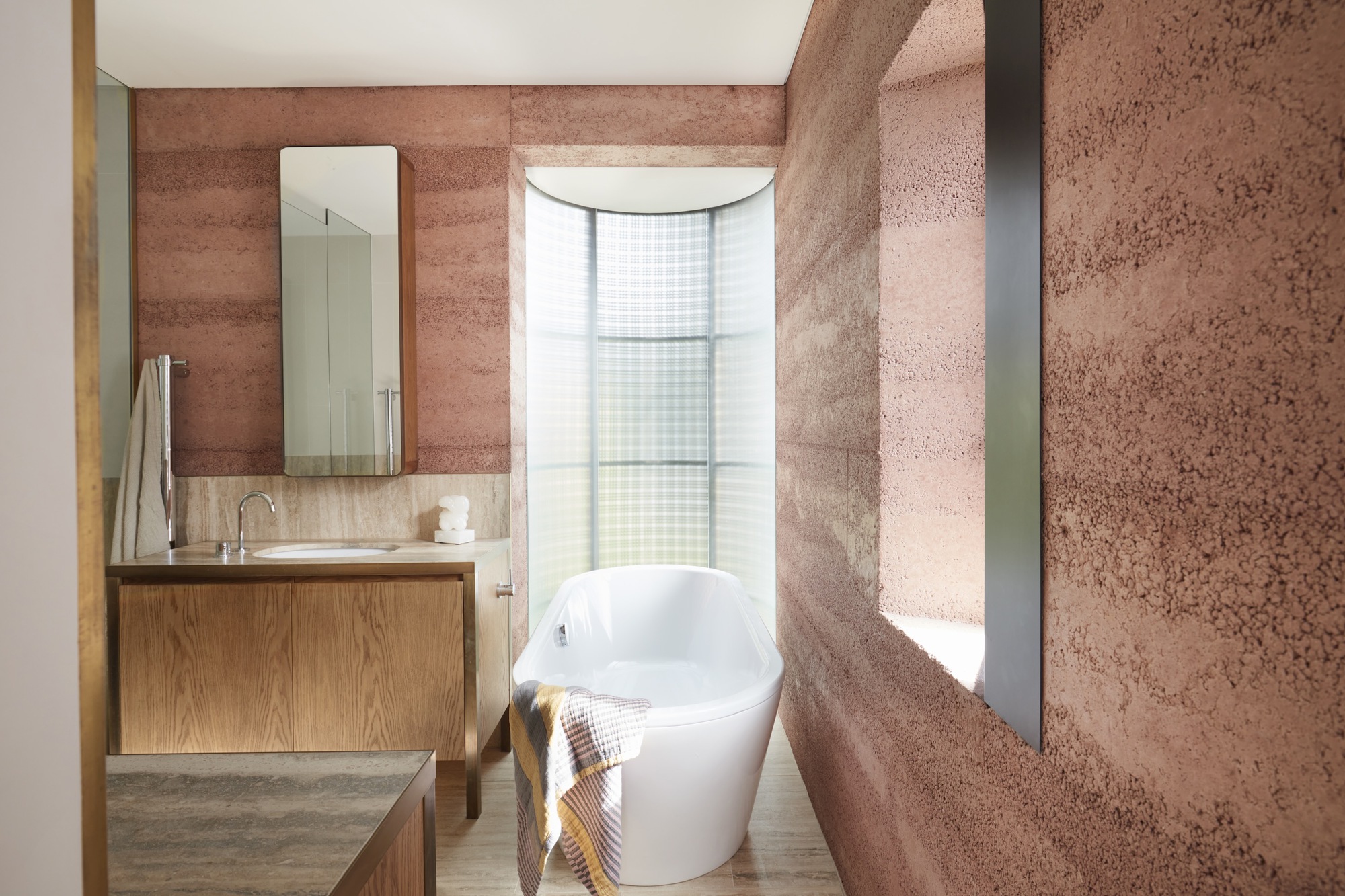Bathroom with rammed earth walls, freestanding bathtub and curved window framing daylight