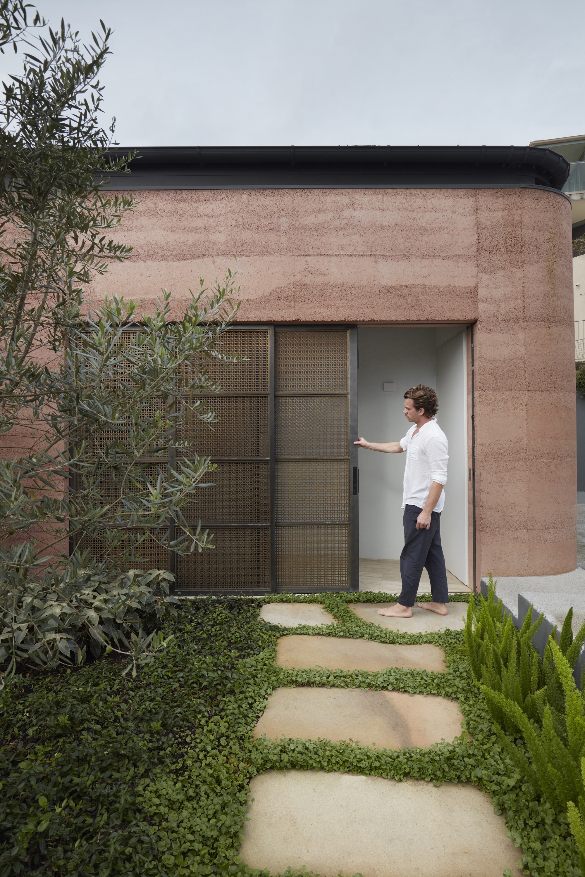 Rammed earth entry facade with perforated metal sliding door and stepping stones through ground cover planting