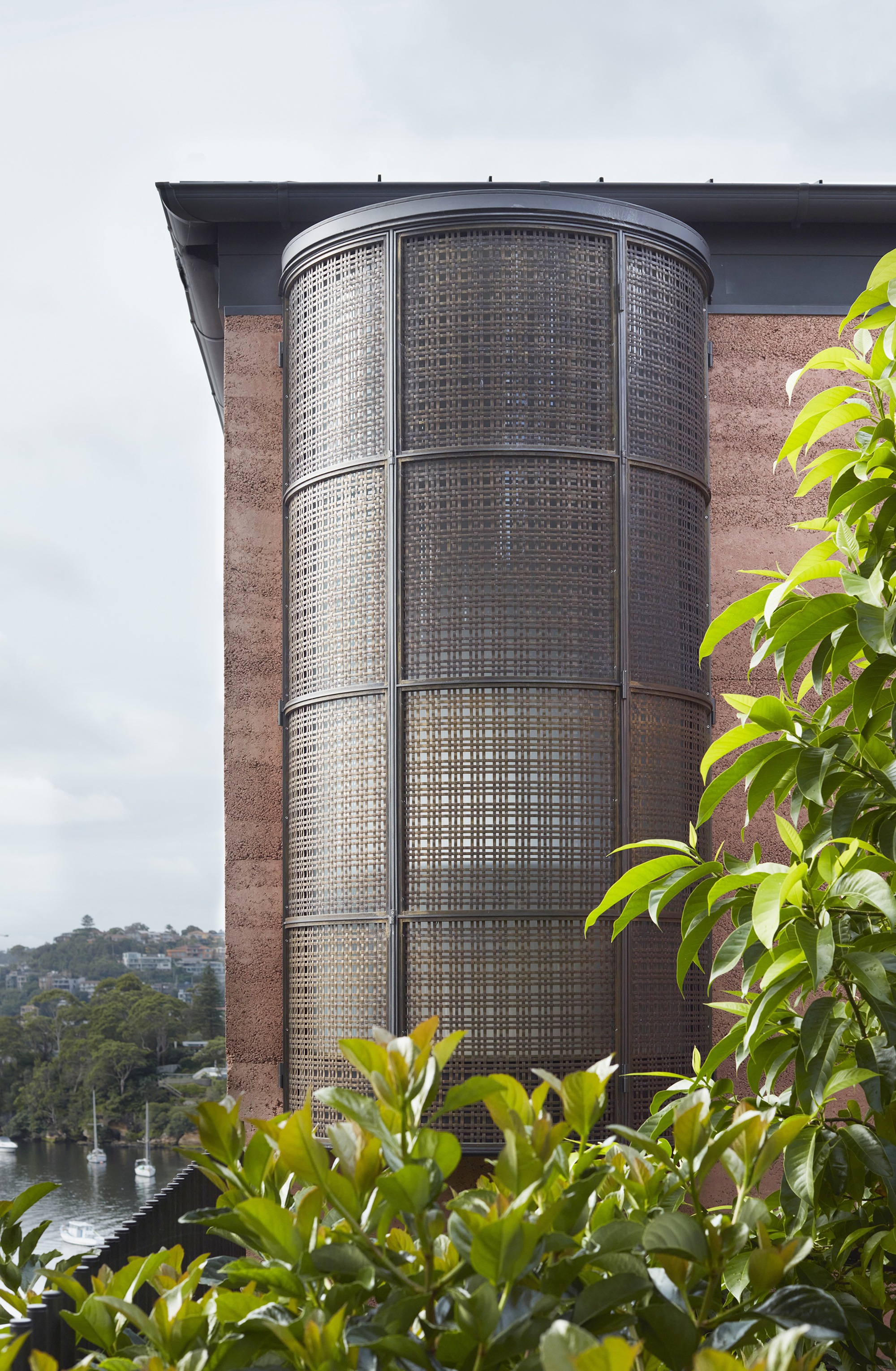 Curved perforated metal screen tower rising against rammed earth wall with hillside visible beyond vegetation