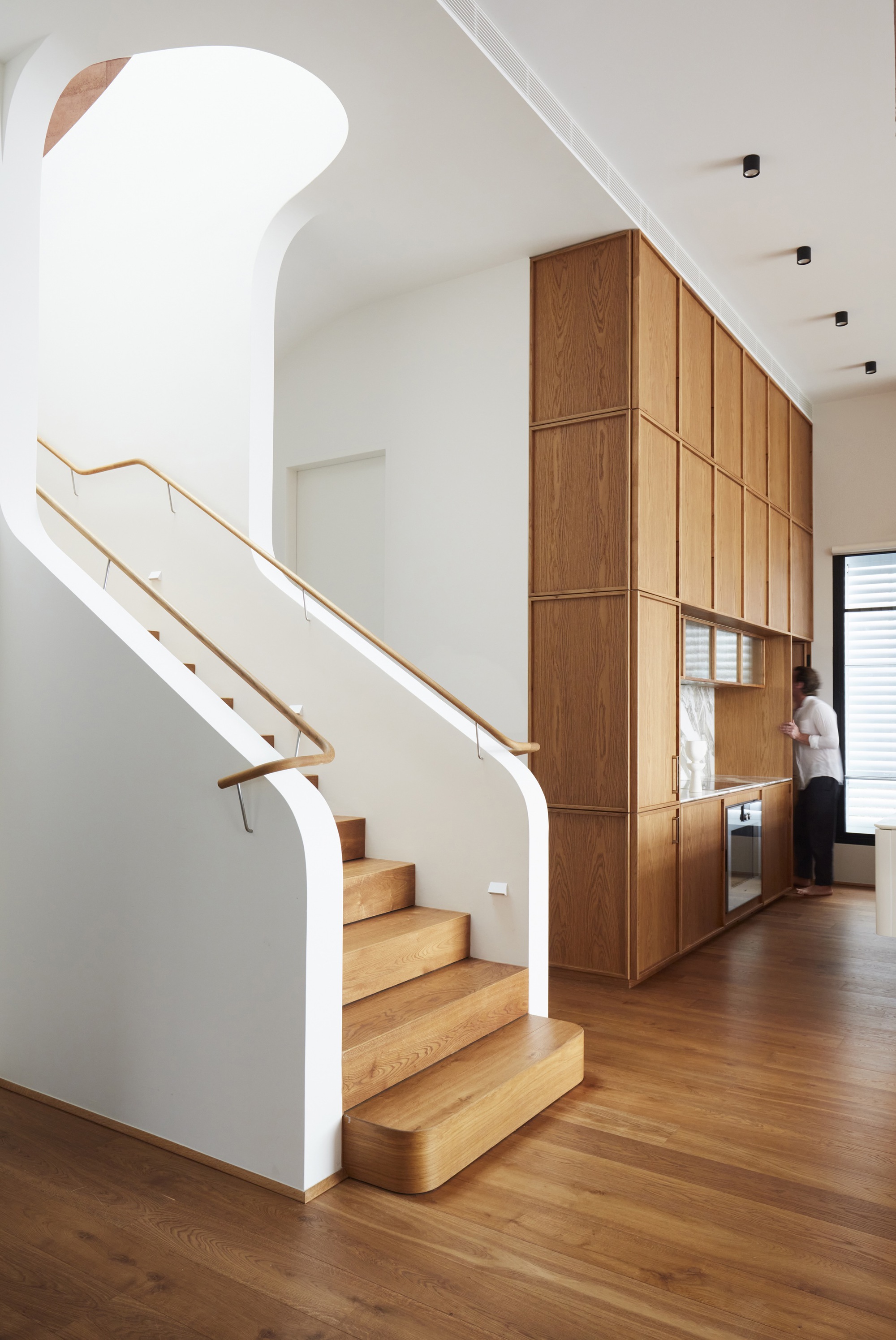 Timber staircase with brass handrail ascending beside full-height oak cabinetry wall with integrated appliances