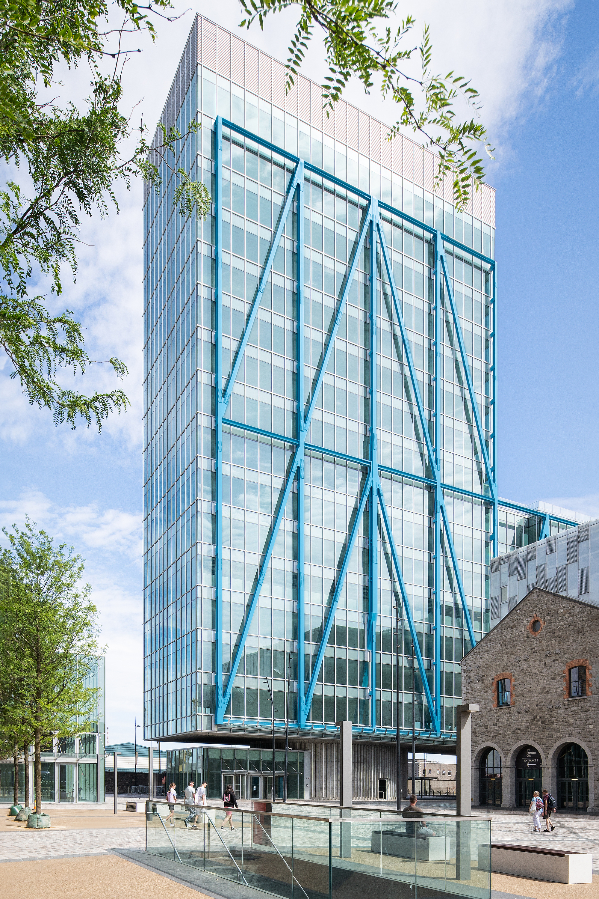 Glass facade with diagonal blue steel bracing framed by tree branches and a public plaza