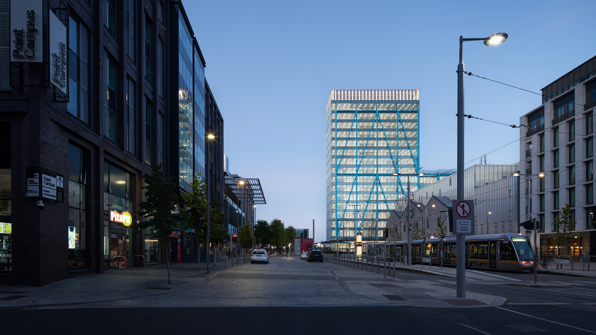 Street view of the illuminated glass and steel tower rising above neighboring buildings at twilight with tram lines