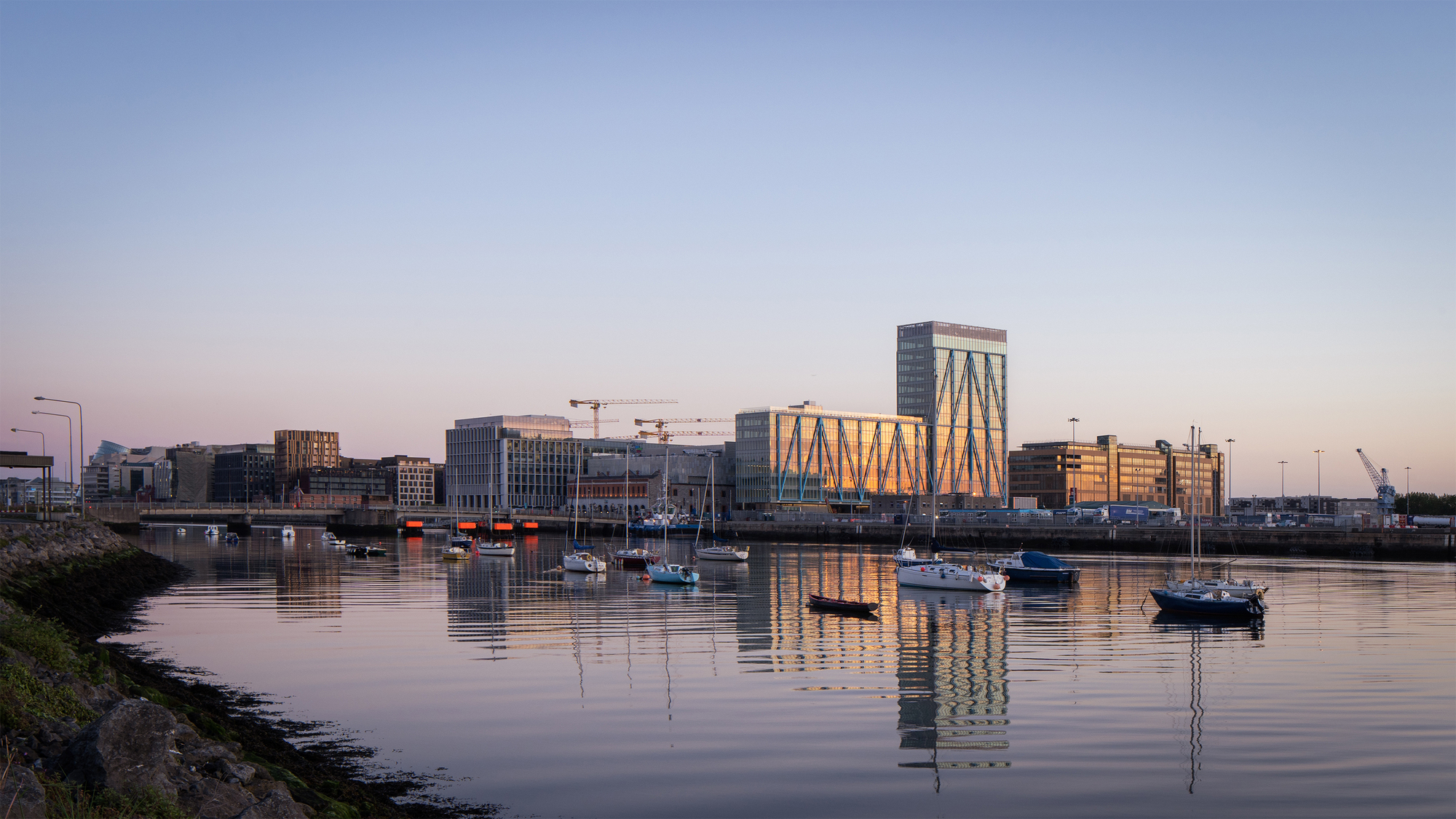 Waterfront view of the diagonal-braced tower reflected in the calm harbor at sunset with moored boats