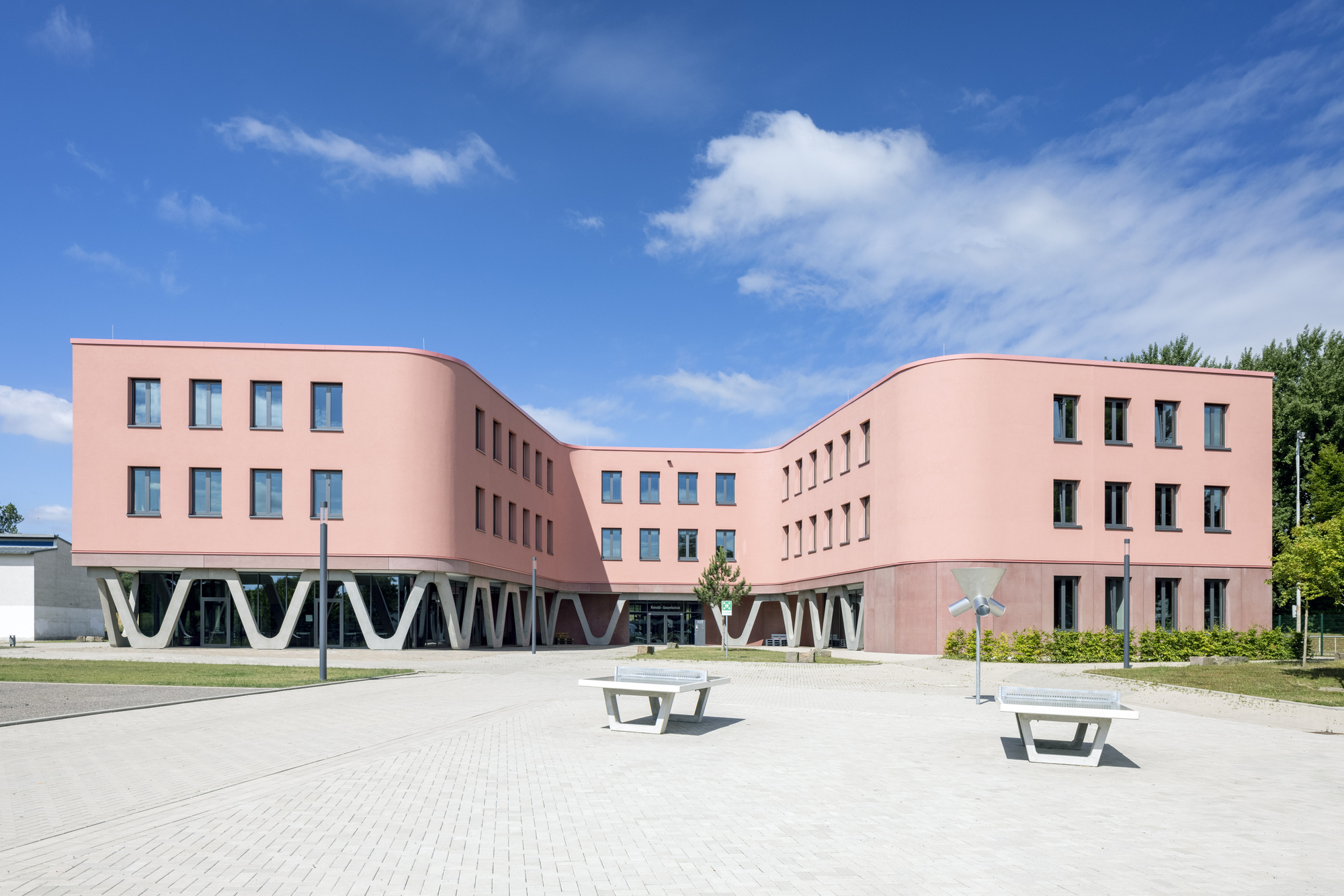 Courtyard view of the pink rendered facade with white V-shaped structural columns and picnic tables