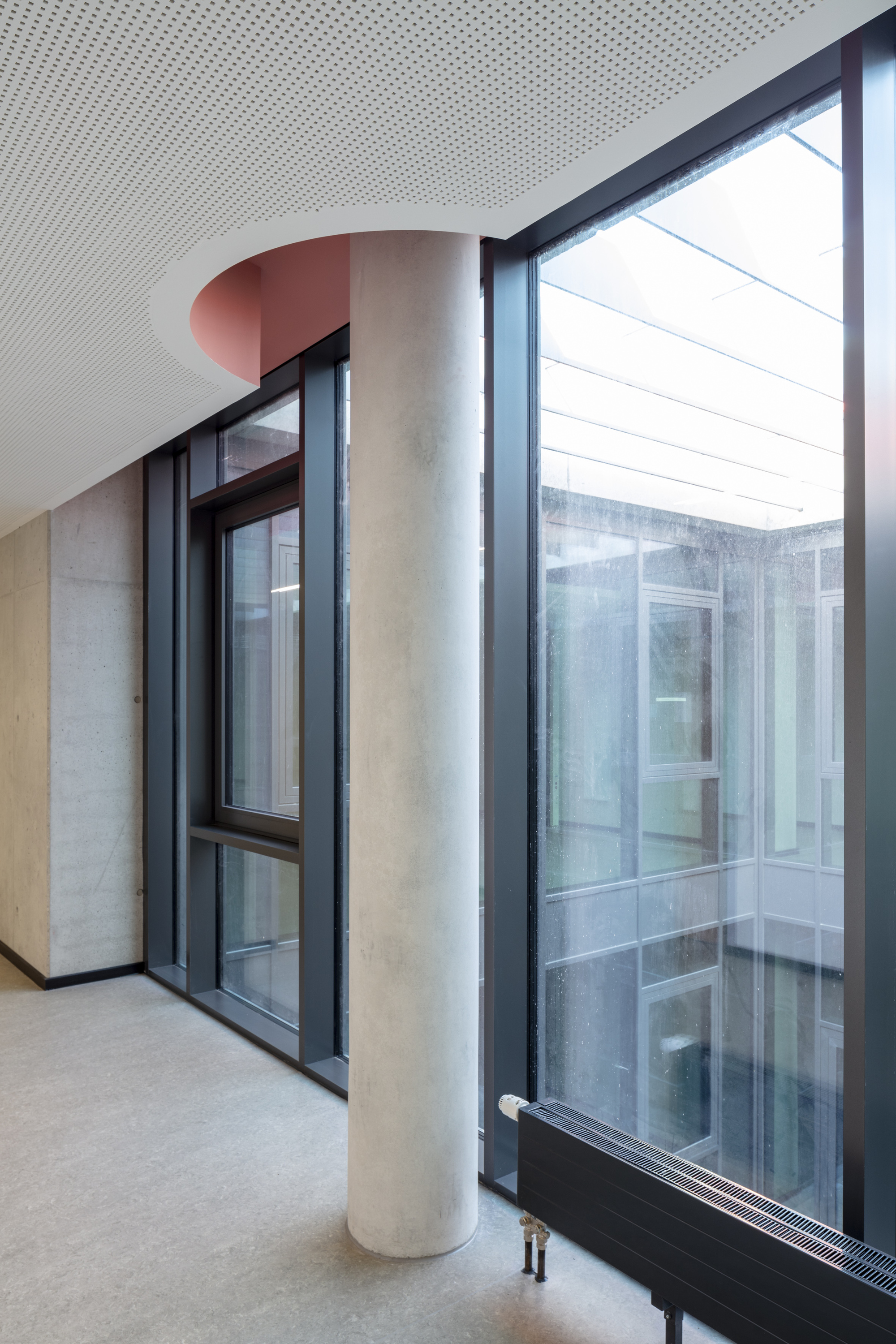Interior corridor with perforated ceiling panel, cylindrical column, and dark-framed glazing overlooking a courtyard