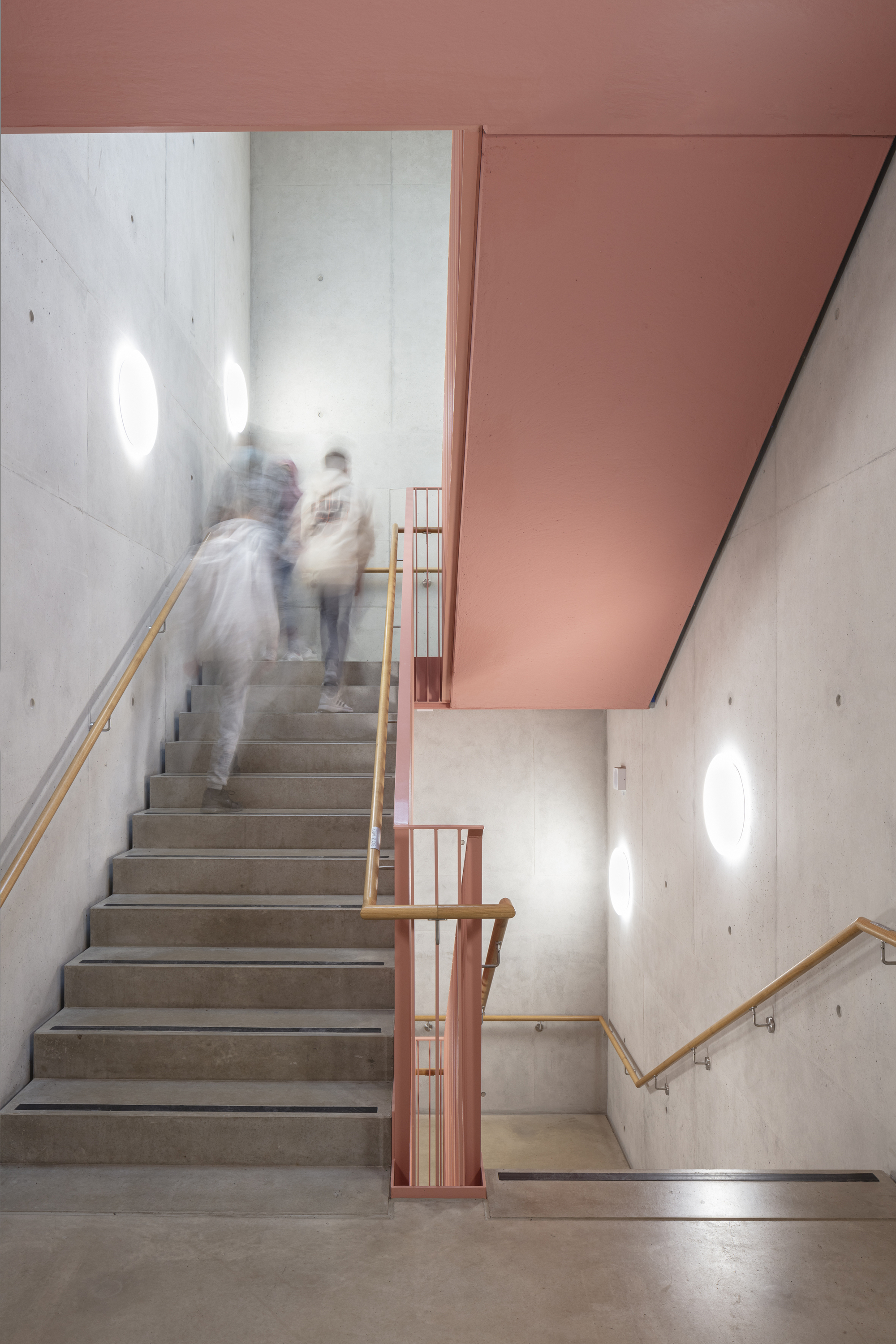 Concrete stairwell with wooden handrails and blurred figures ascending beneath a pink soffit