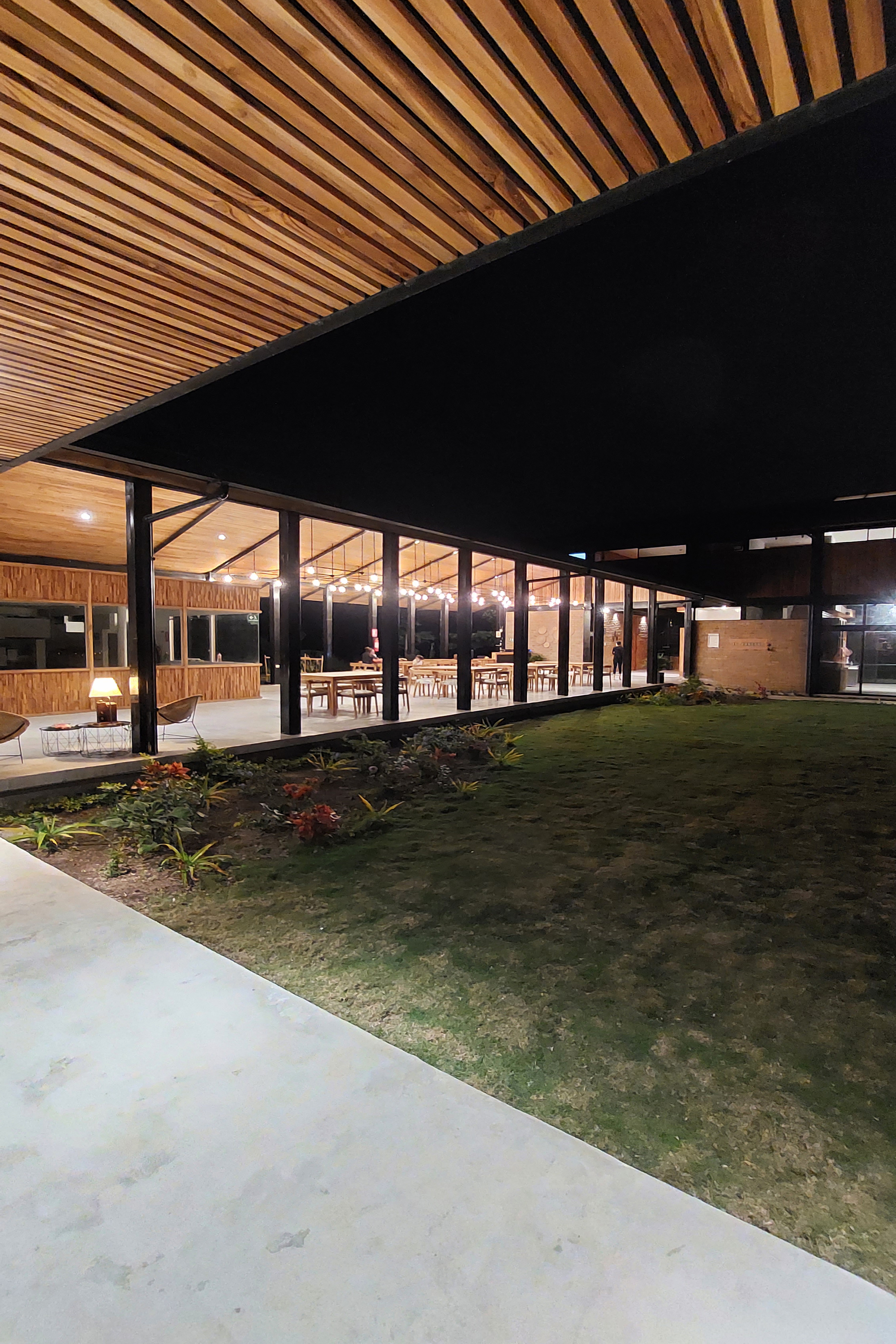 Covered outdoor terrace with timber slatted ceiling, steel columns, and a central planted courtyard at dusk