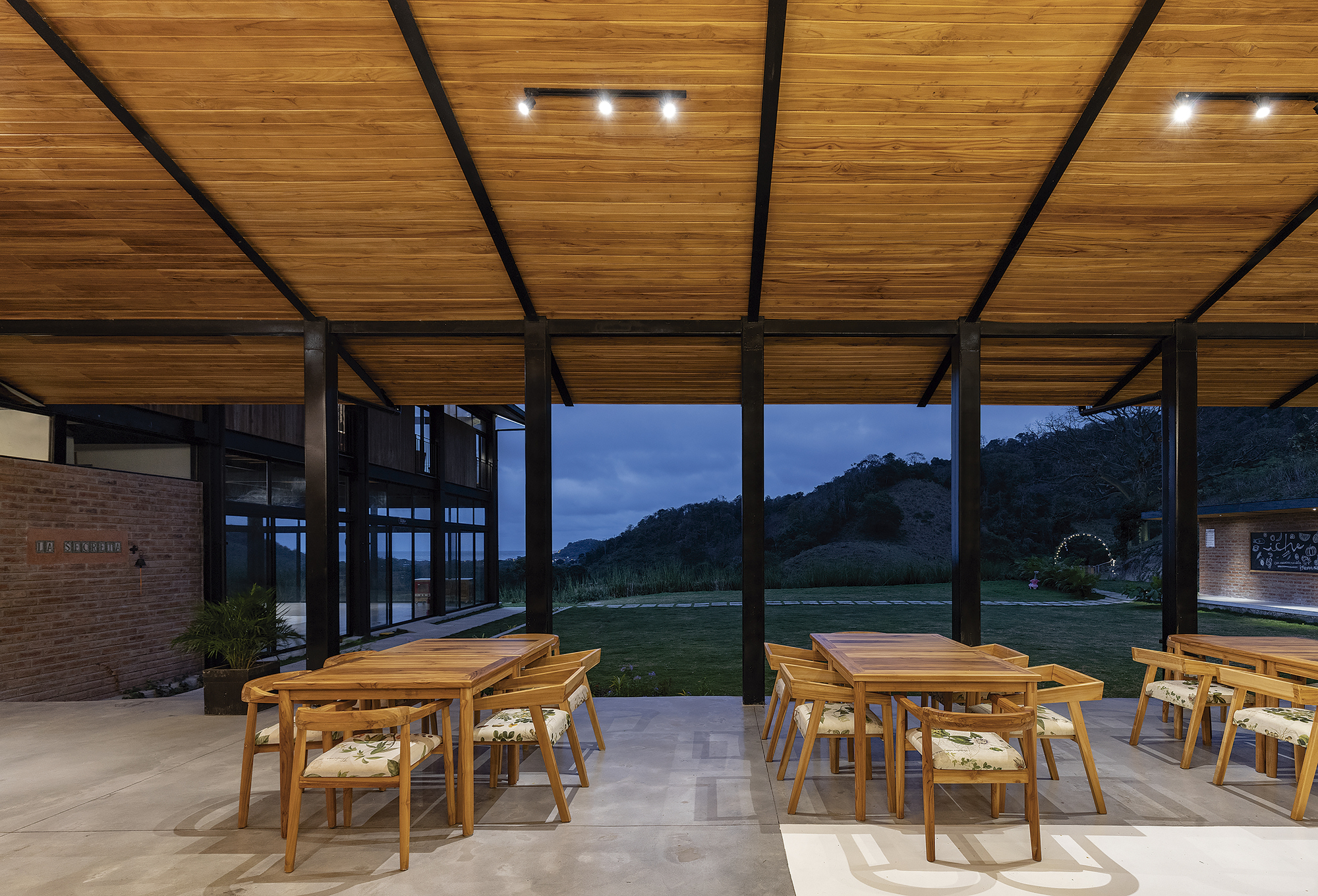 Open-air dining terrace with timber ceiling panels and steel beams overlooking hills at dusk