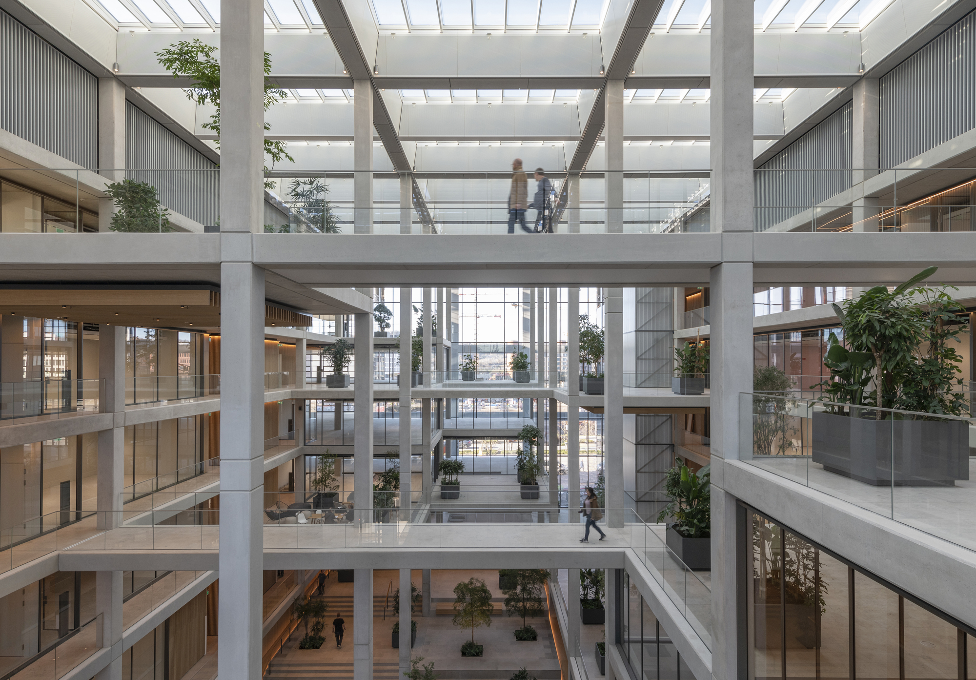 Multi-storey atrium with concrete beams, glass skylight and people crossing a central bridge
