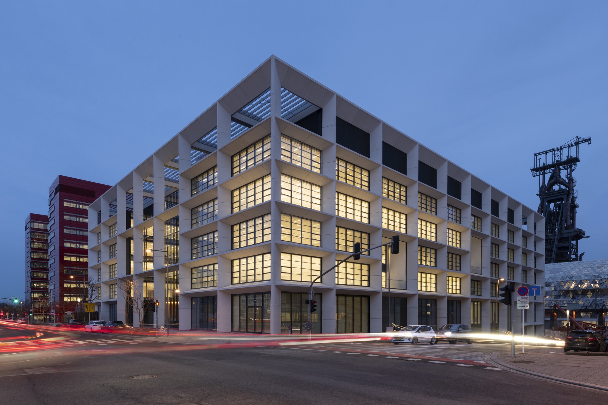 Street corner view at dusk showing the gridded facade with an industrial tower visible beyond