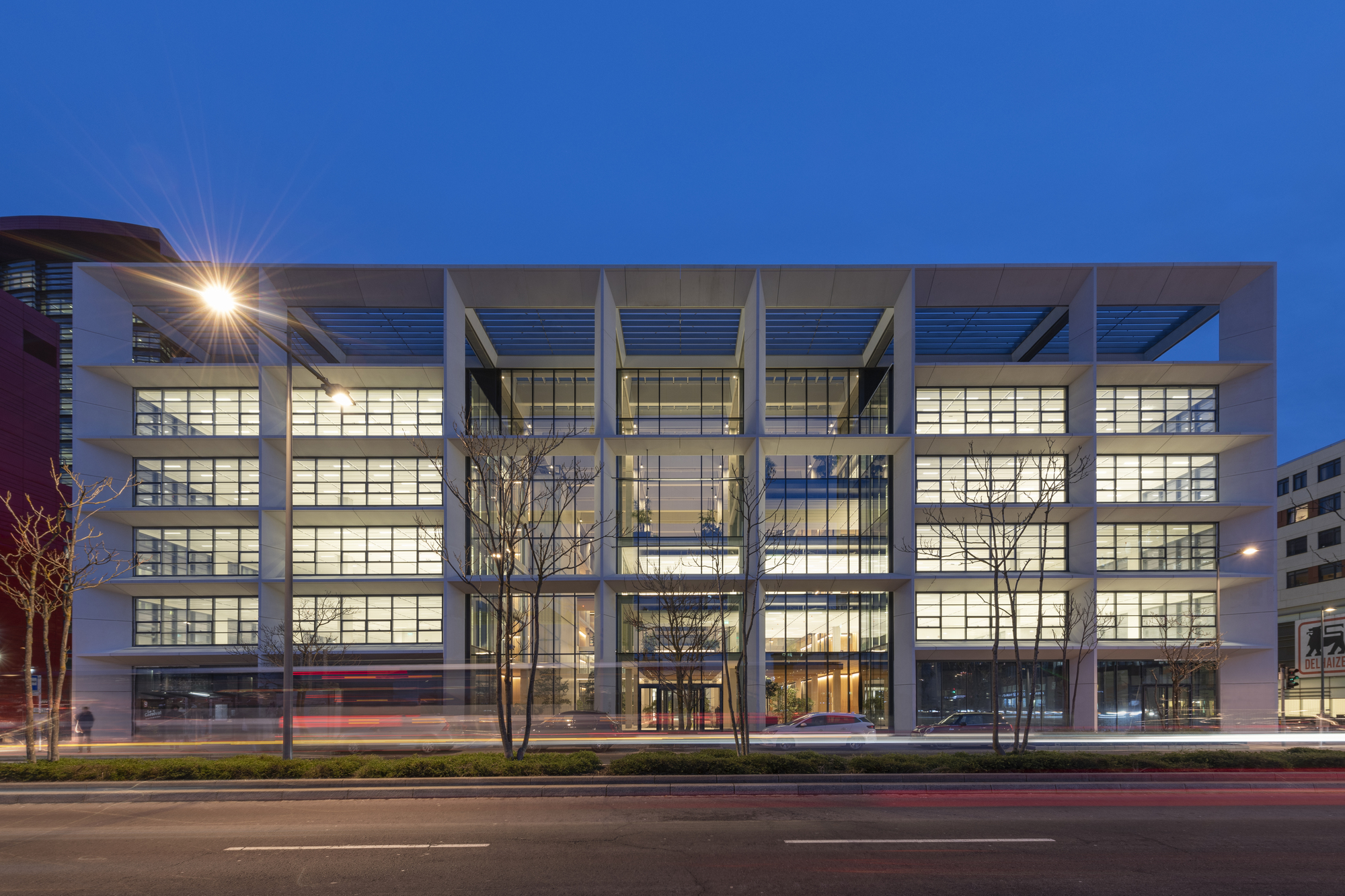 Front facade at twilight with illuminated glazing framed by deep concrete beams and bare trees