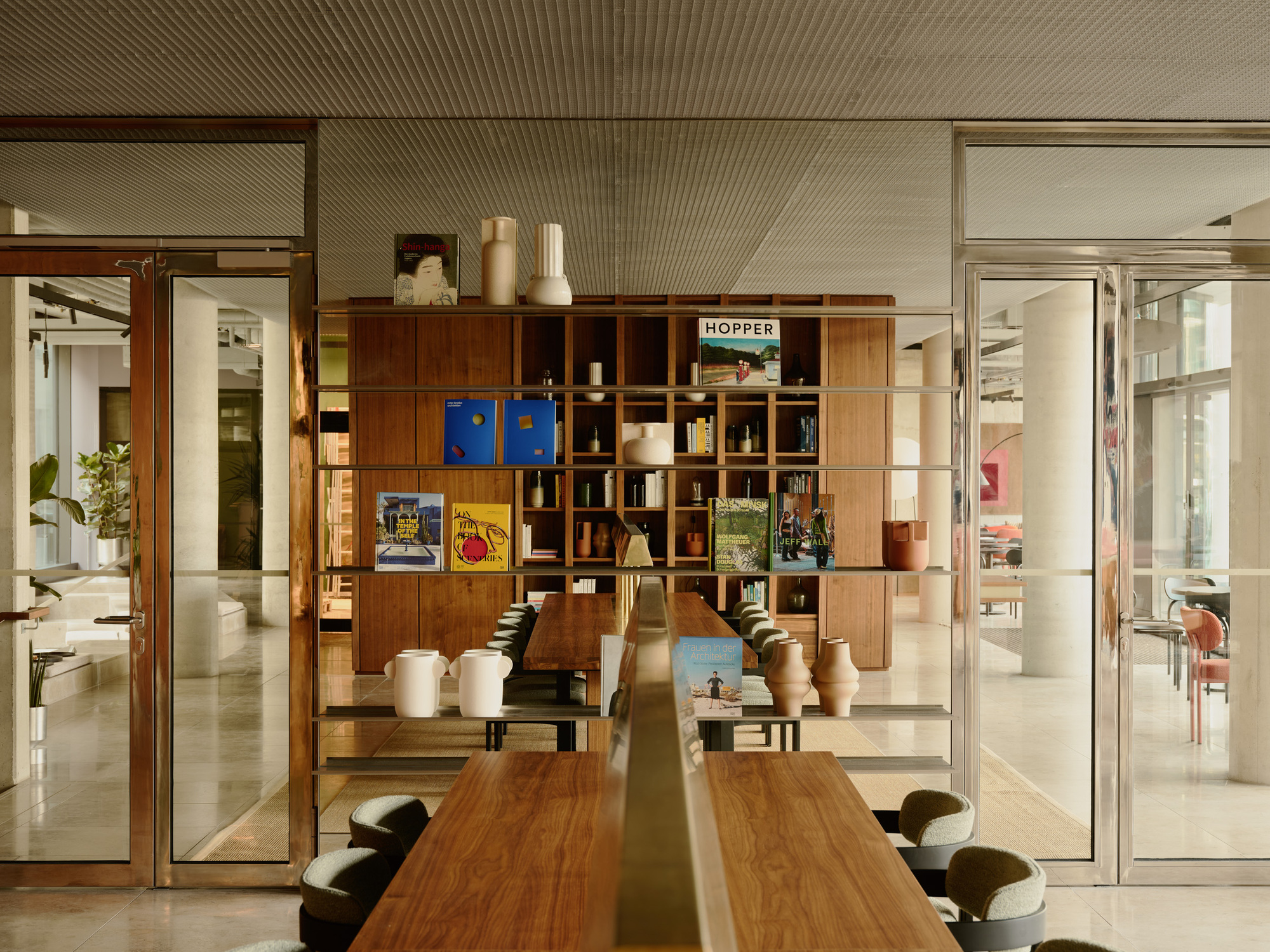 Walnut dining table facing a timber shelving wall with glass doors opening to adjacent office spaces
