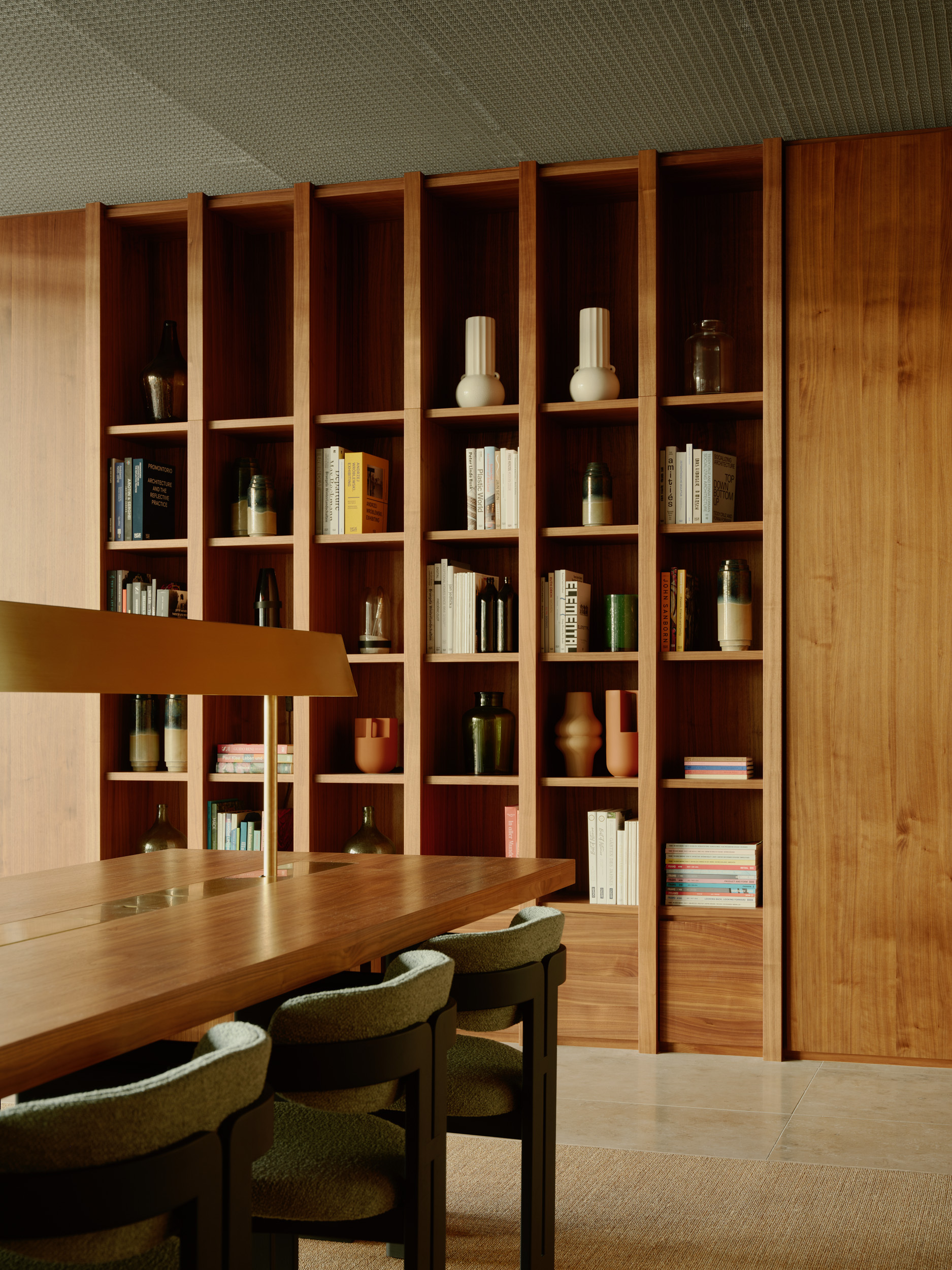 Timber shelving wall with ceramic objects and books beside dining table with green upholstered chairs