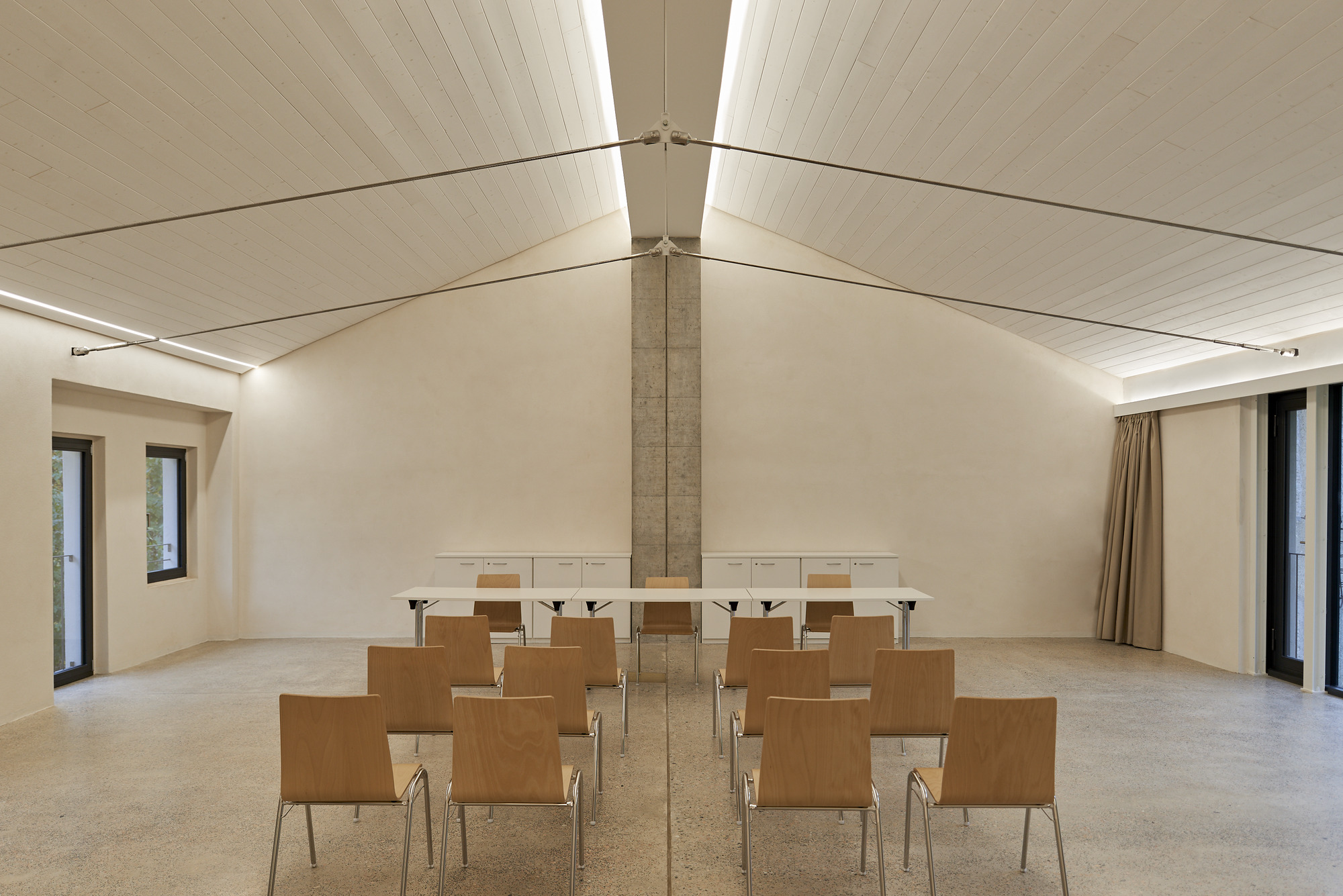 Meeting room with vaulted timber ceiling, central concrete column and rows of plywood chairs on terrazzo floor