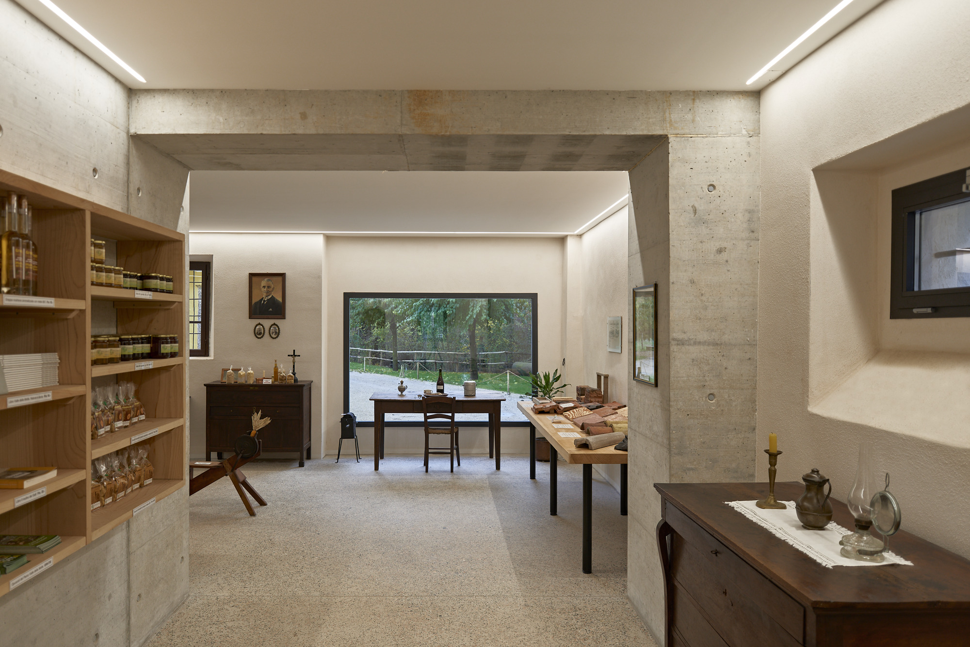 View through concrete portal frame to exhibition room with timber shelving, dining table and garden beyond glass