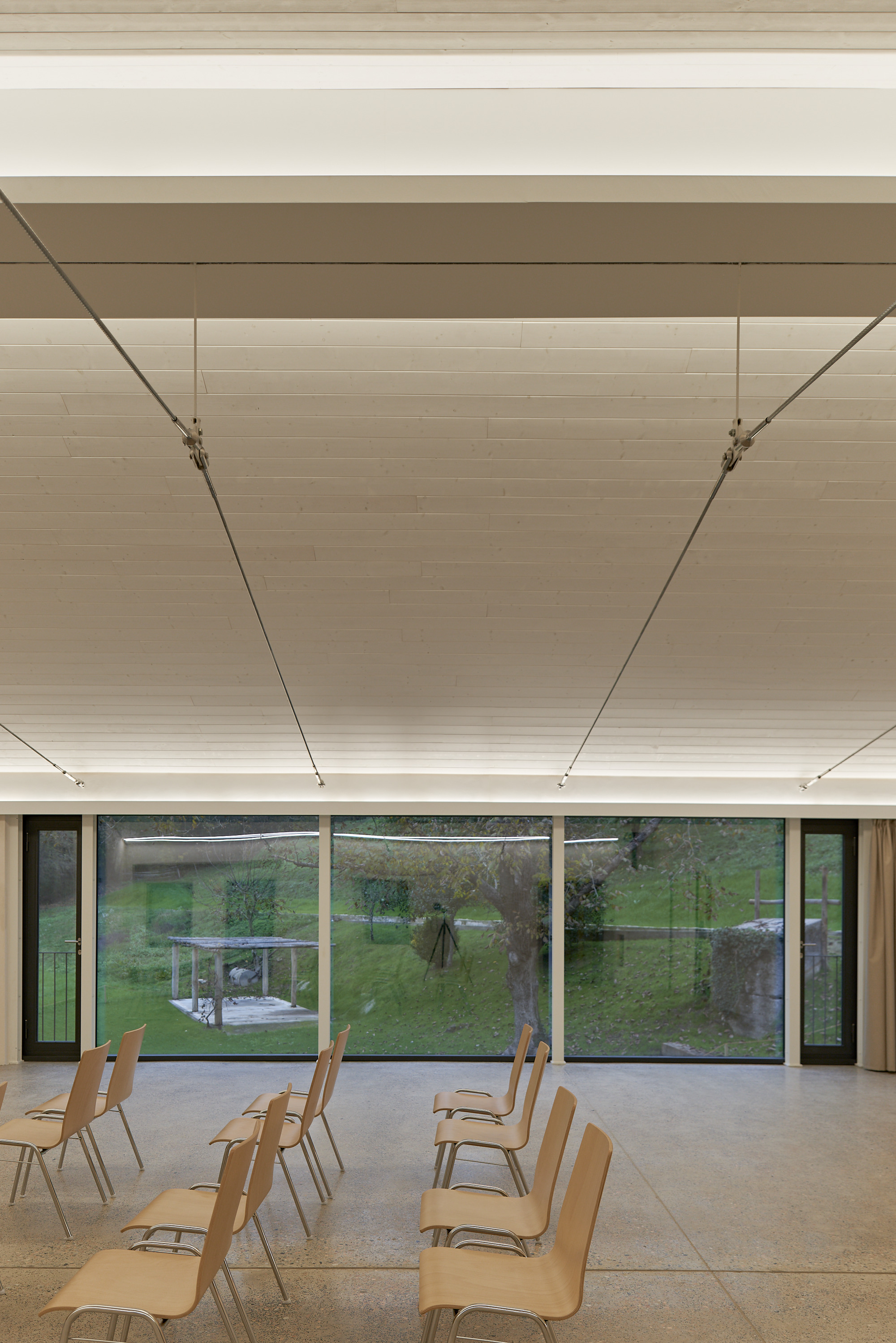 Interior multipurpose room with tensioned fabric ceiling panels, plywood chairs, and glazed wall overlooking garden