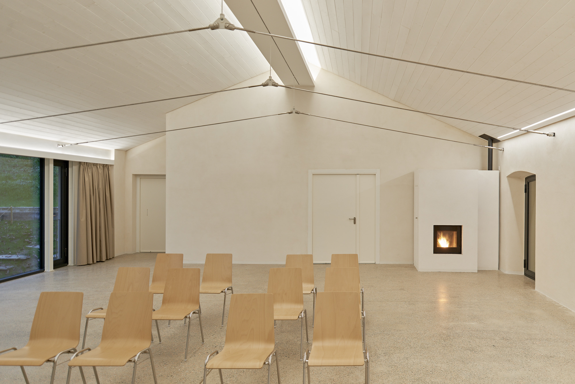 Assembly space with gabled fabric ceiling, terrazzo floor, plywood seating, and freestanding fireplace with active flame