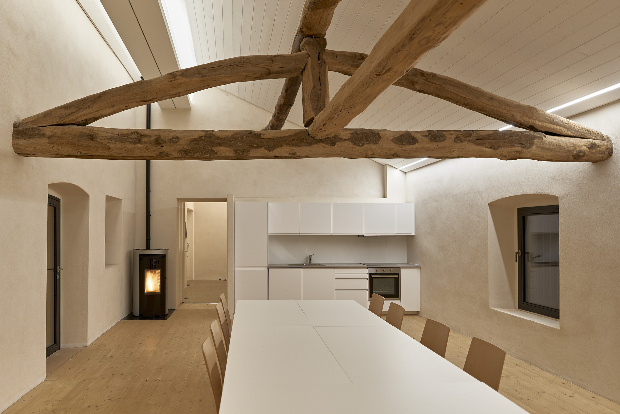 Dining area with exposed timber roof truss above white table and kitchen cabinetry along rear wall
