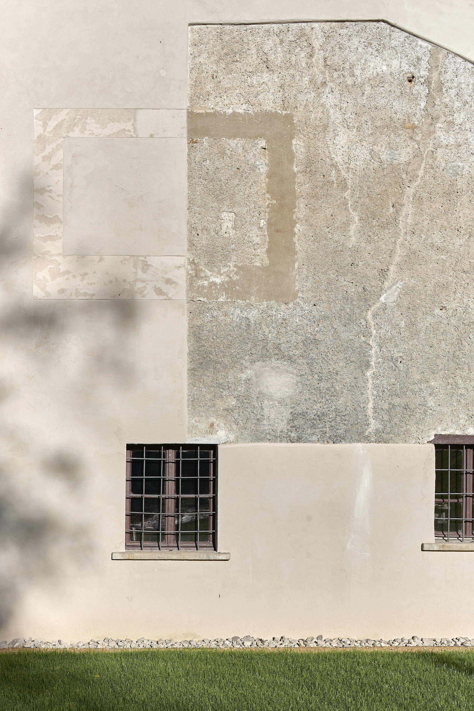 Layered plaster facade with patched repairs and two barred windows above a grass lawn