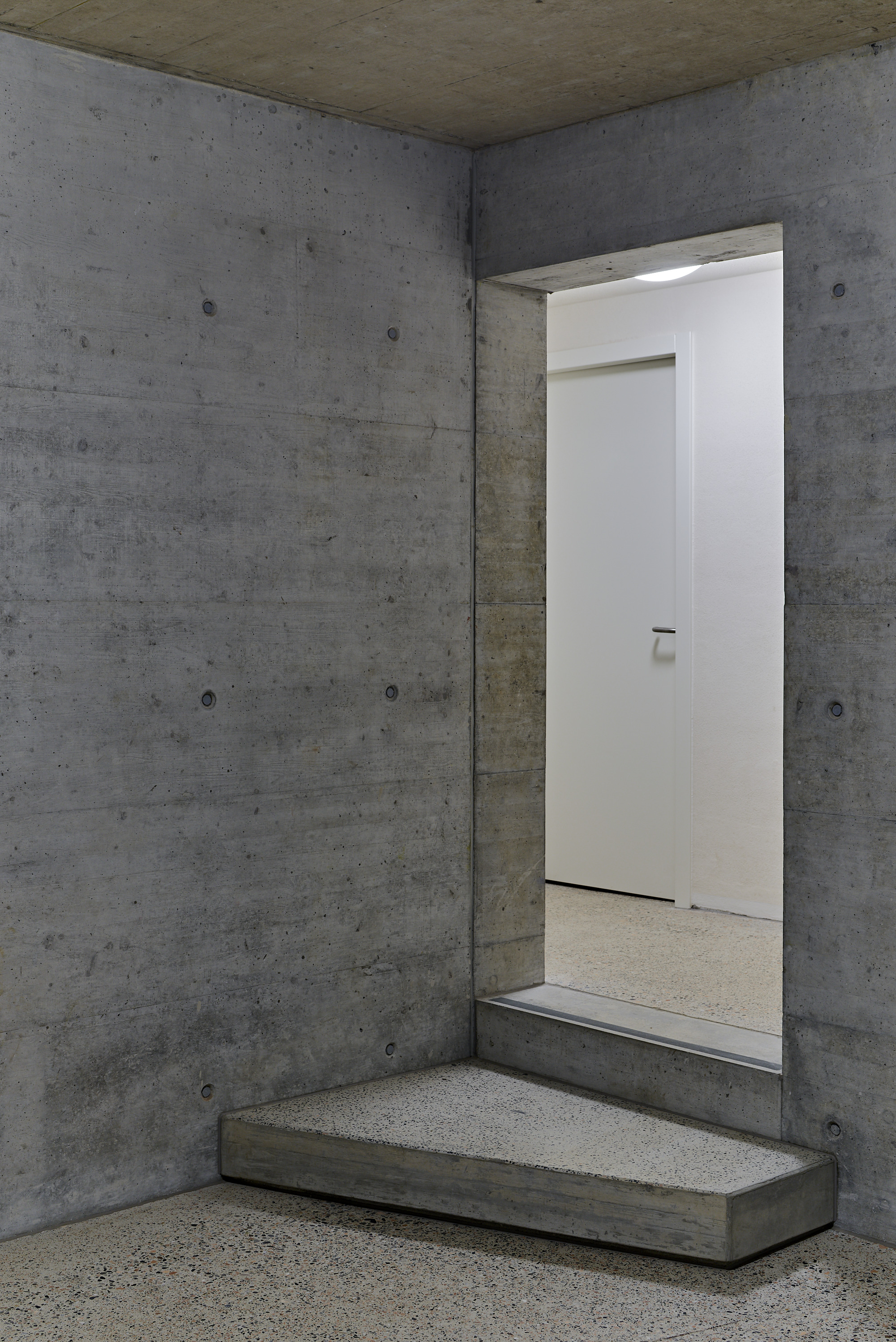 Board-formed concrete entryway with stepped platform and white door opening in dim interior light