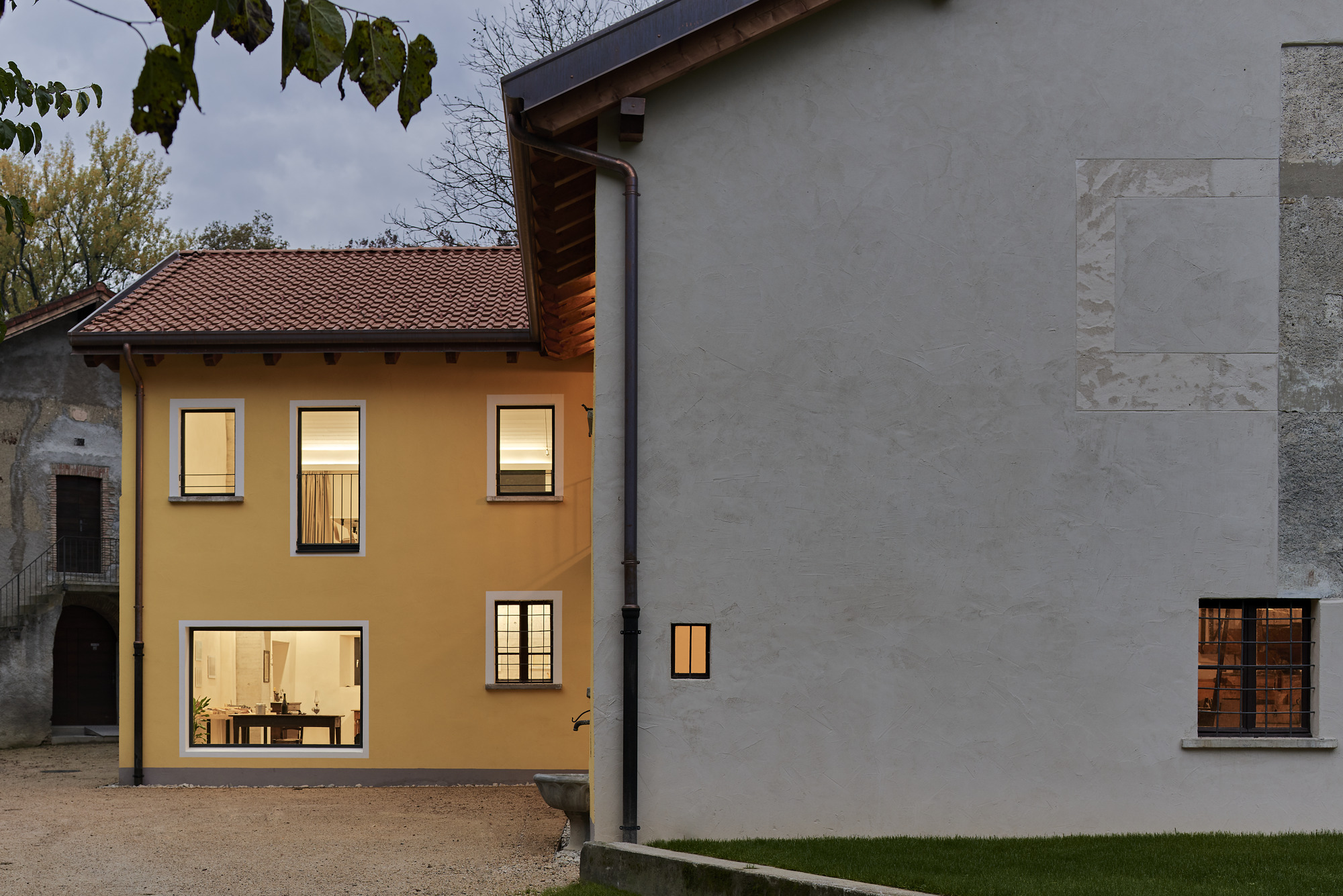 Courtyard view of illuminated yellow house and grey rendered addition at dusk