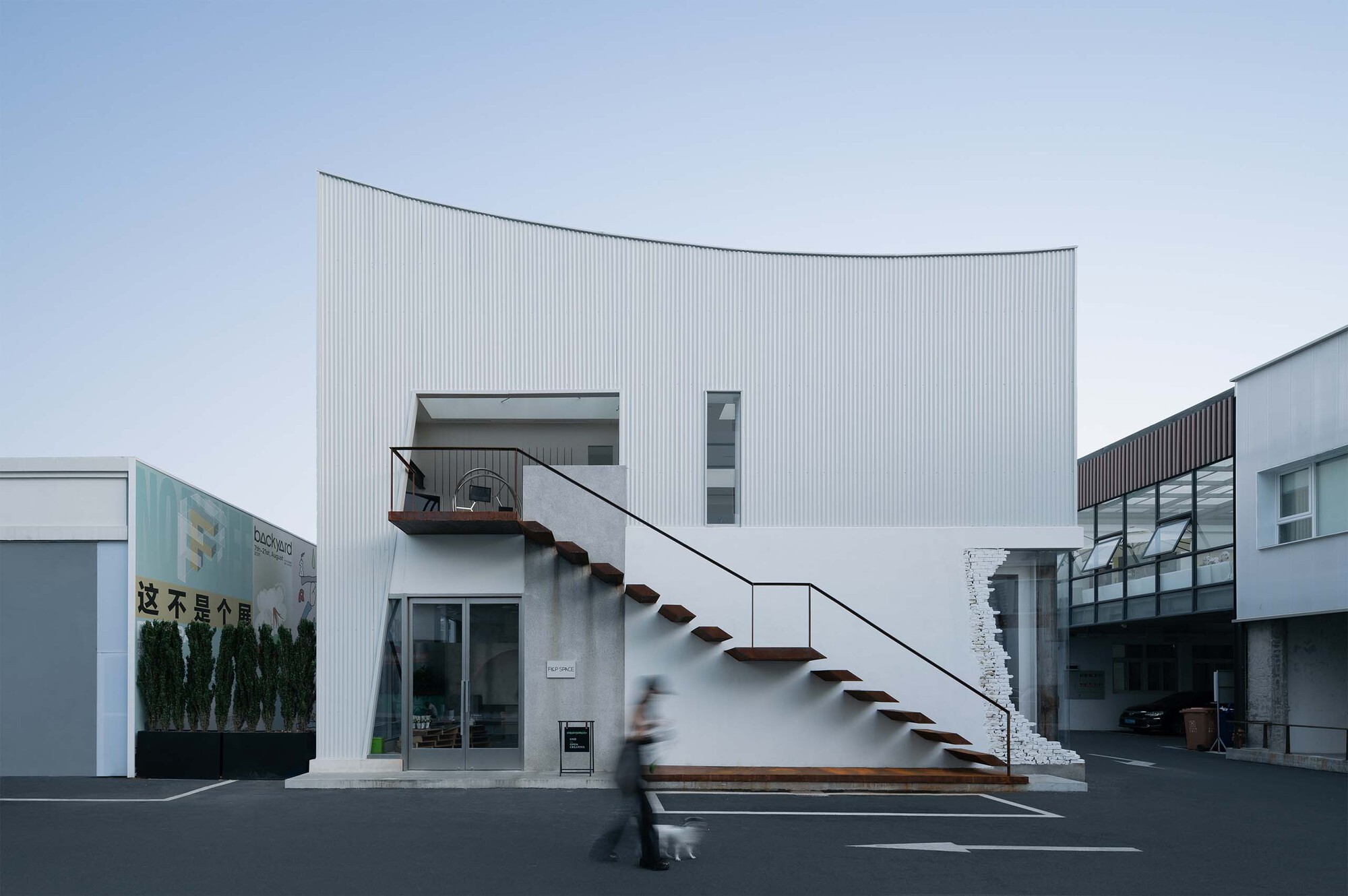 External staircase with open timber treads ascending to the curved corrugated facade as a figure walks below